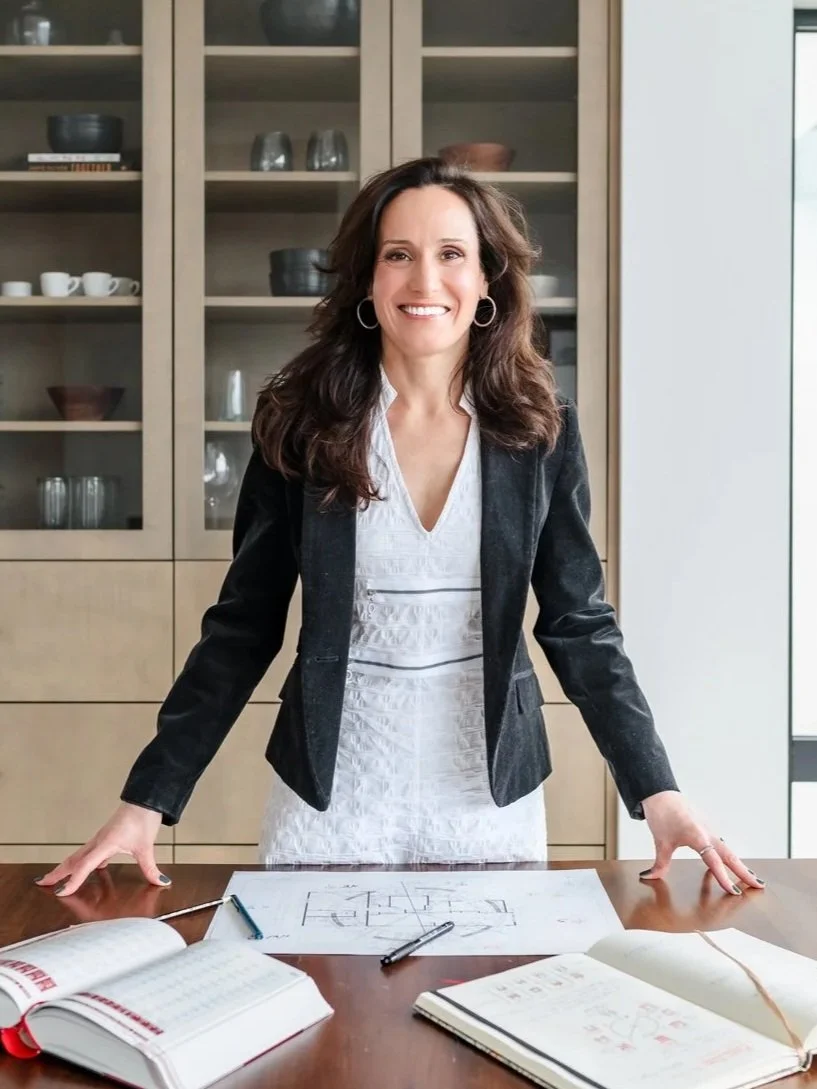 Elena in a white dress and black blazer standing behind a desk with architectural plans, Chinese Metaphysics almanac, pens, and a notebook.
