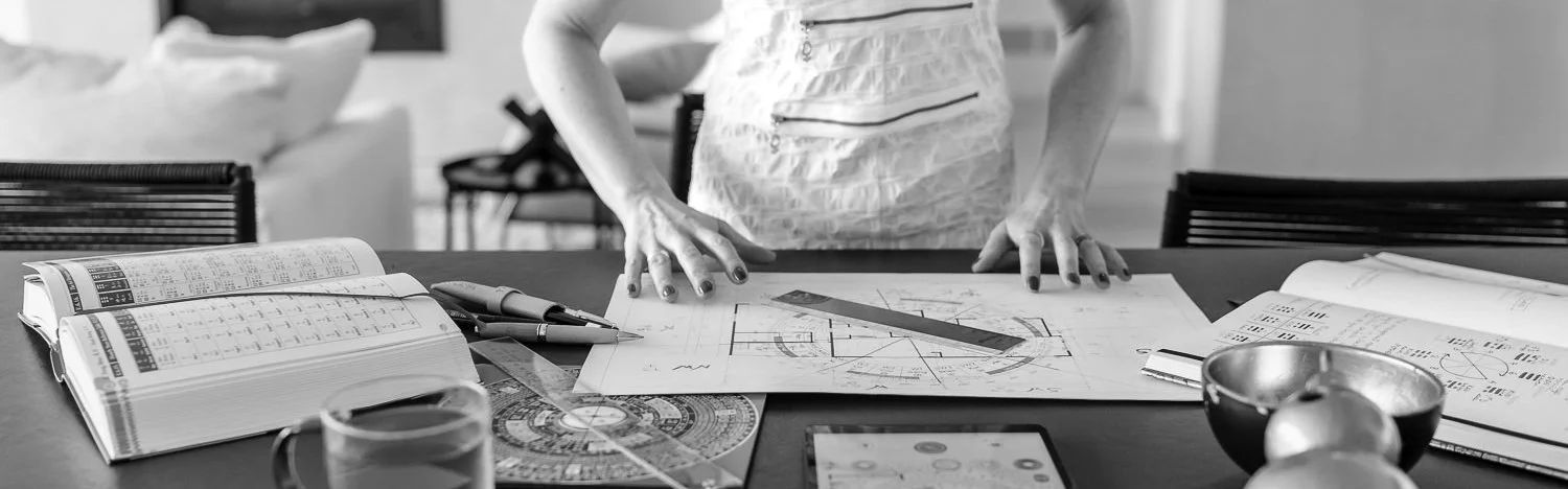 Woman working at a desk with architectural blueprints, measuring tools, a Luo Pan compass and Feng Shui books.