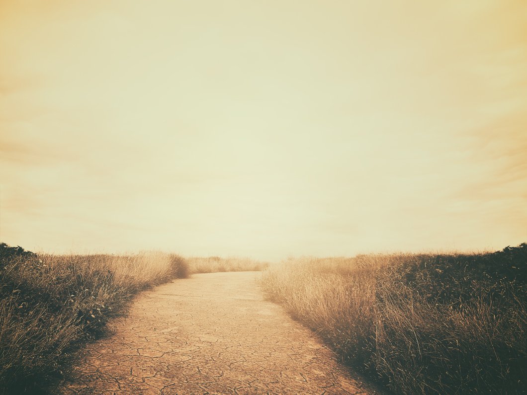 A path in the countryside winding through grass on both sides with a bright sun in the sky.