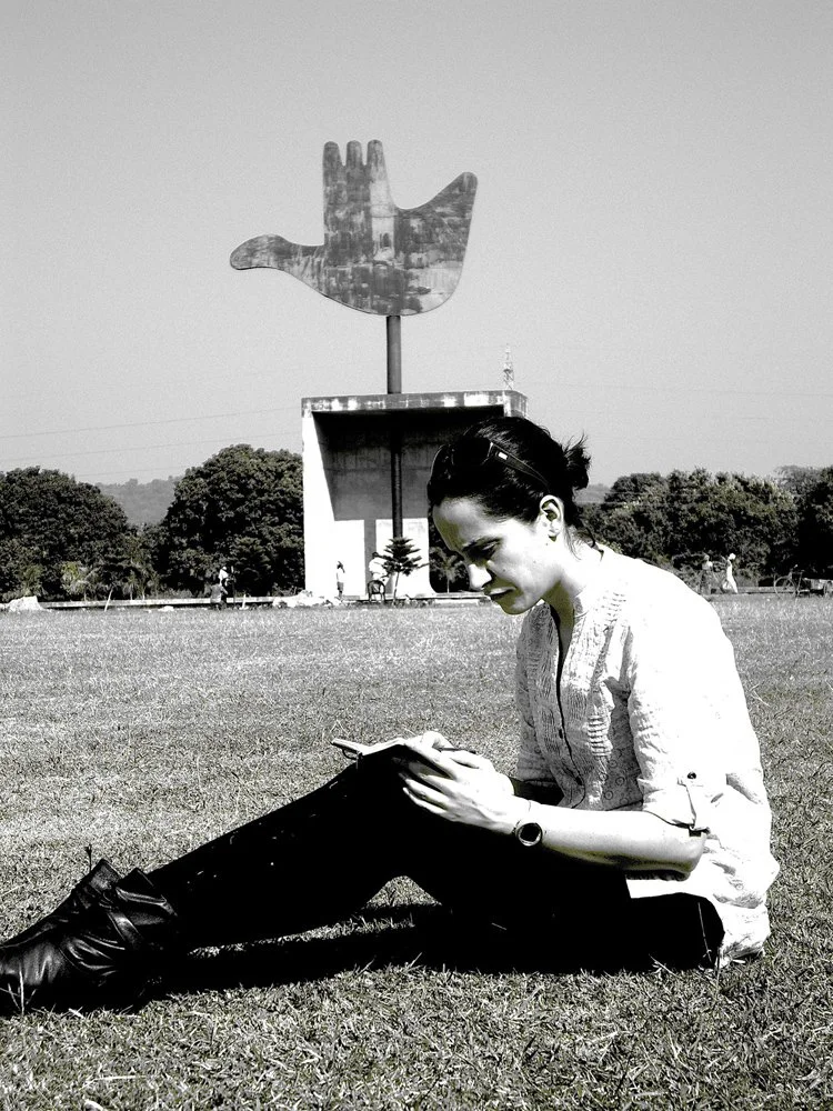 Elena sitting on the grass, reading a book, in front of the Open Hand monument designed by Le Corbusier in Chandigarh.