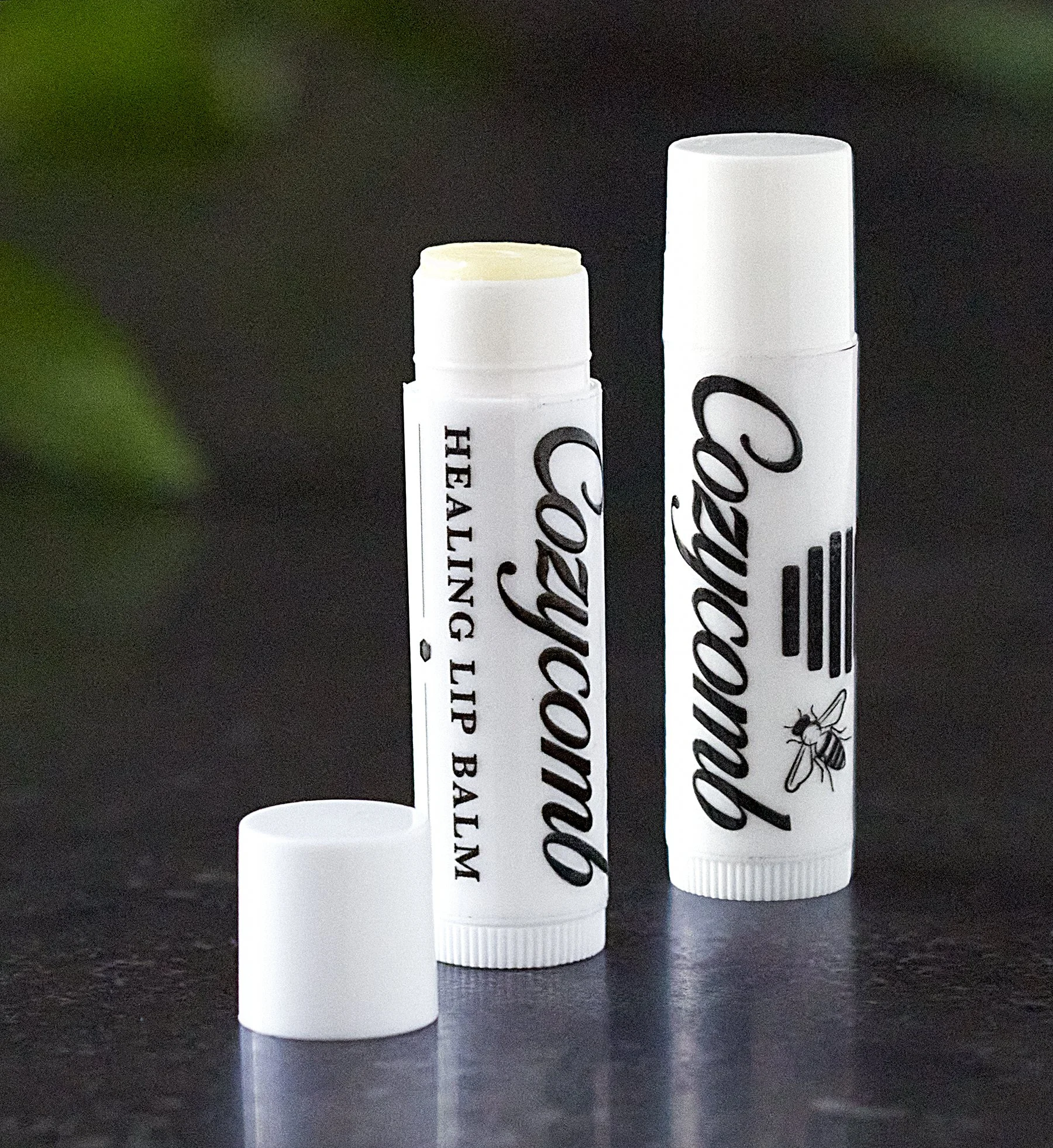 A white lip balm tube labeled 'Coconut' and 'Healing Lip Balm' with the cap removed, placed on a dark surface with a blurred green background.