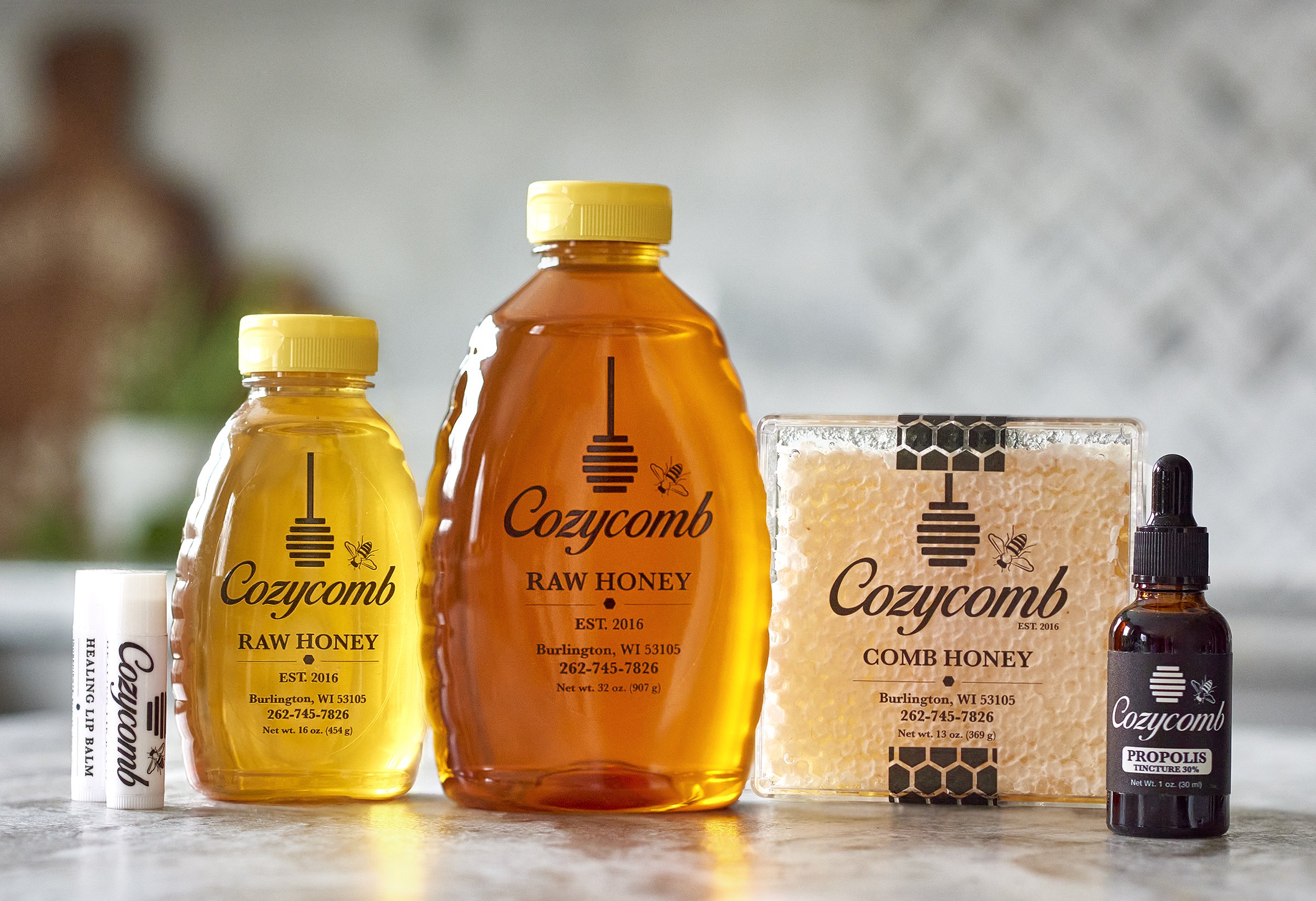 Assorted Cozycomb honey products including raw honey bottles, comb honey, lip balm, and propolis tincture on a kitchen counter.