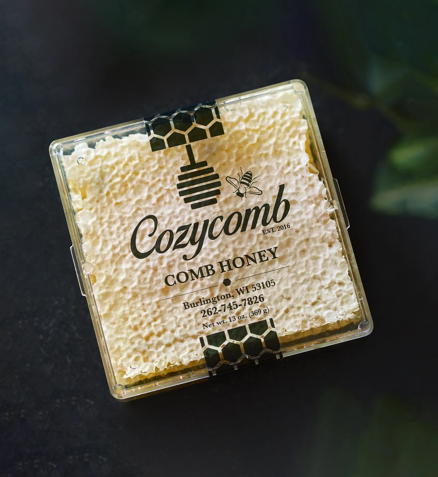 Square container of Cozycomb comb honey with black and honeycomb design on lid, label reads 'Cozycomb', 'Comb Honey', with location Burlington, WI 53105 and phone number 262-745-7826.