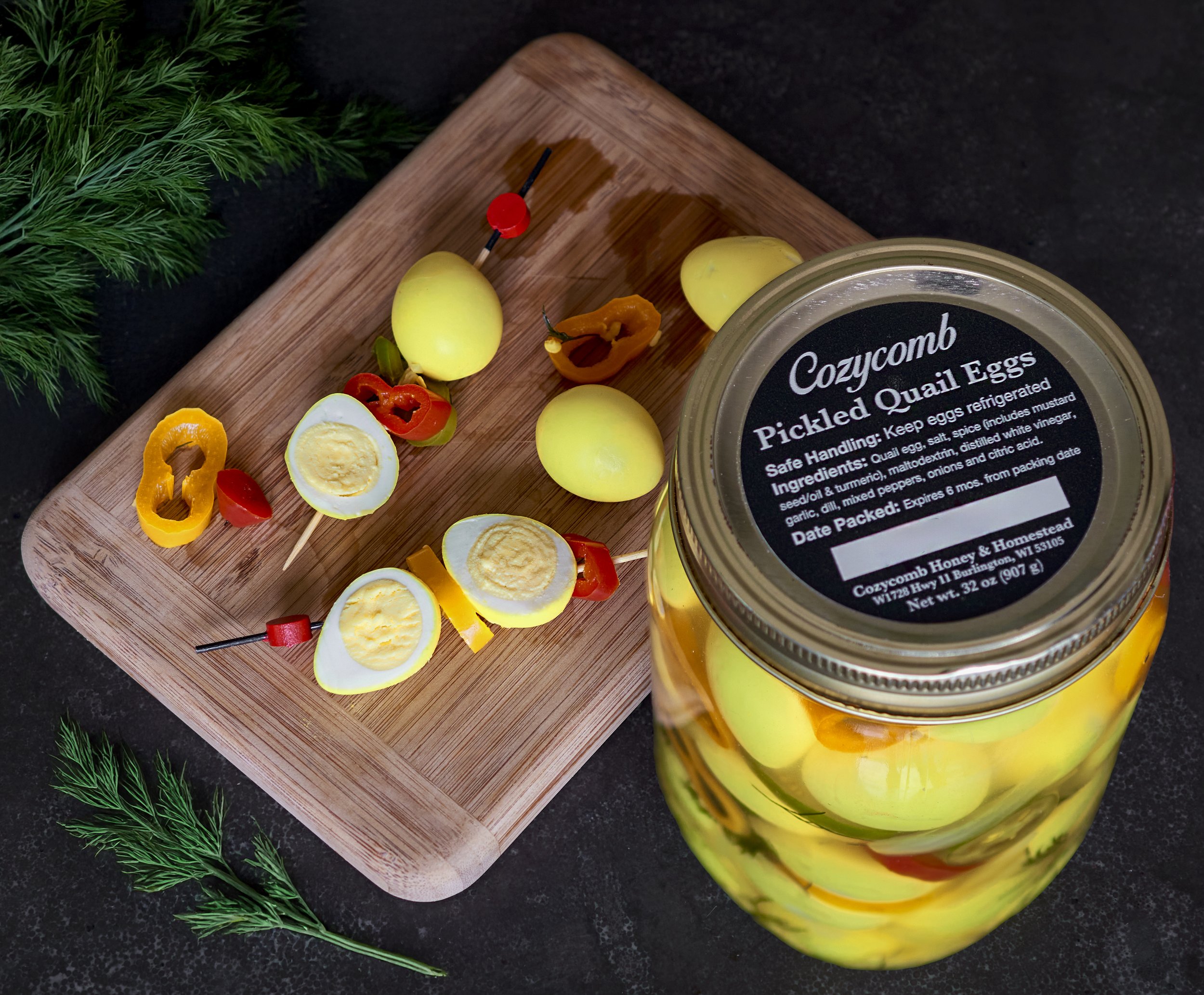 A jar of cozt comb pickled quail eggs on a black countertop, with some eggs and pepper skewers on a wooden cutting board nearby, decorated with green dill herbs.
