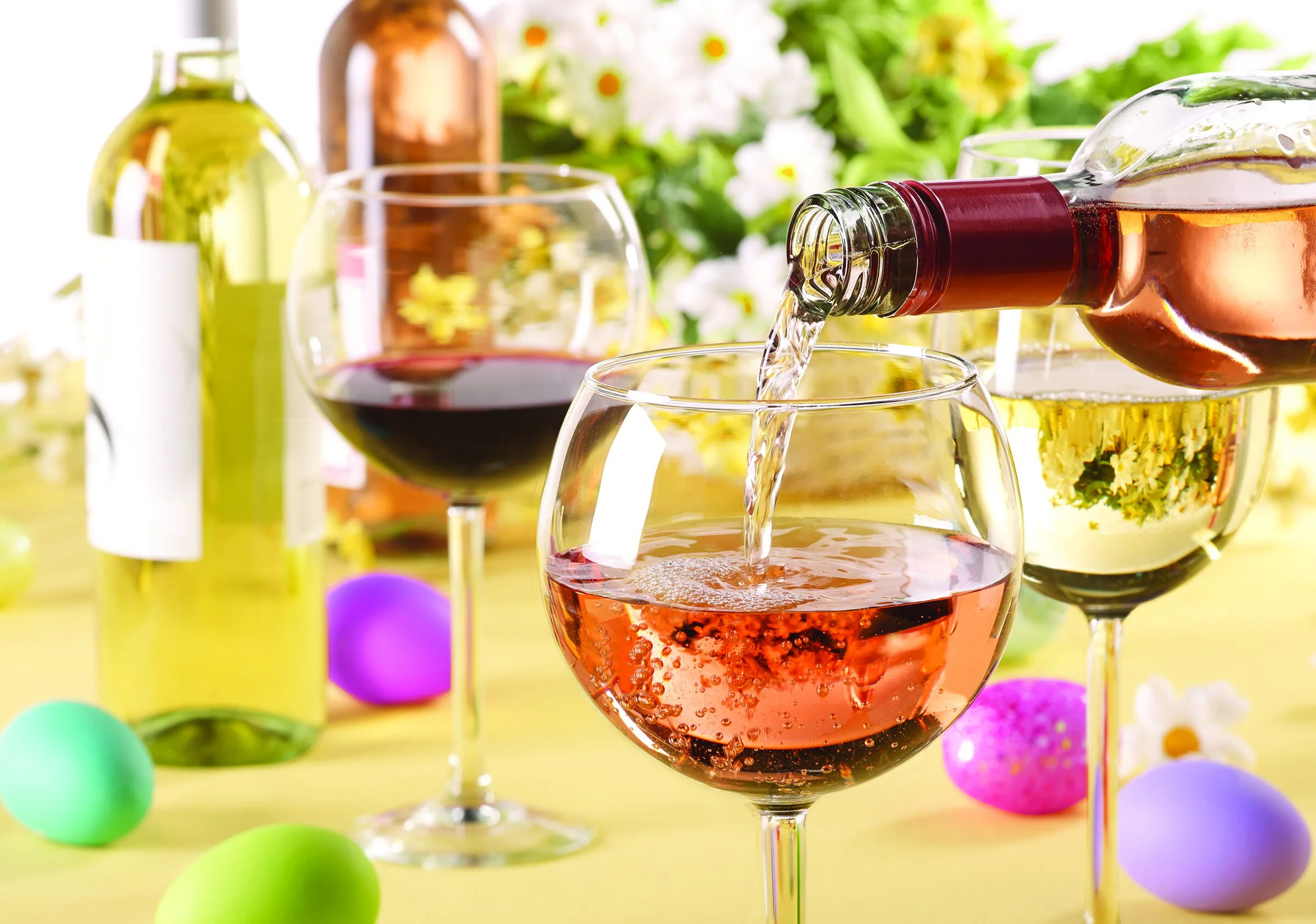 Pouring rosé wine into a wine glass at a party with multiple wine bottles, glasses of red and white wine, colorful eggs, and floral decorations on a yellow tablecloth.