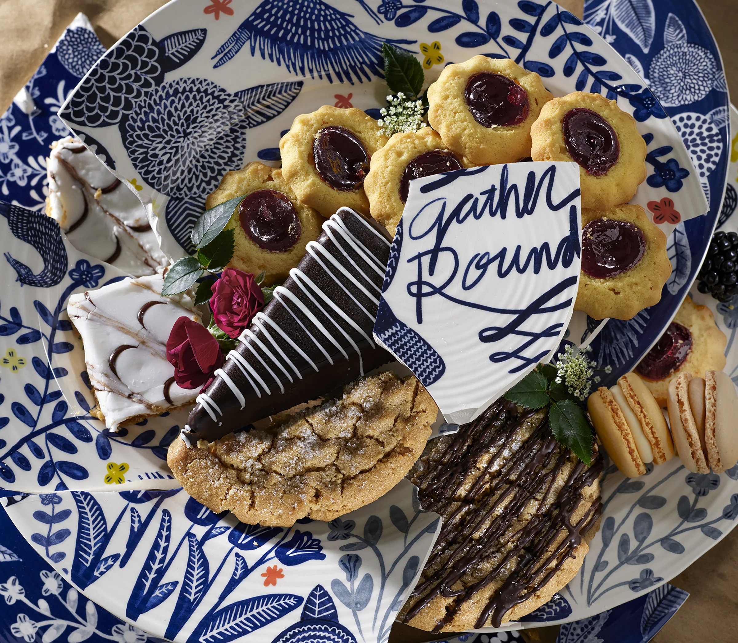 Assorted cookies, macarons, and chocolates on a decorative blue and white plate with a card that says 'Gather Round'.
