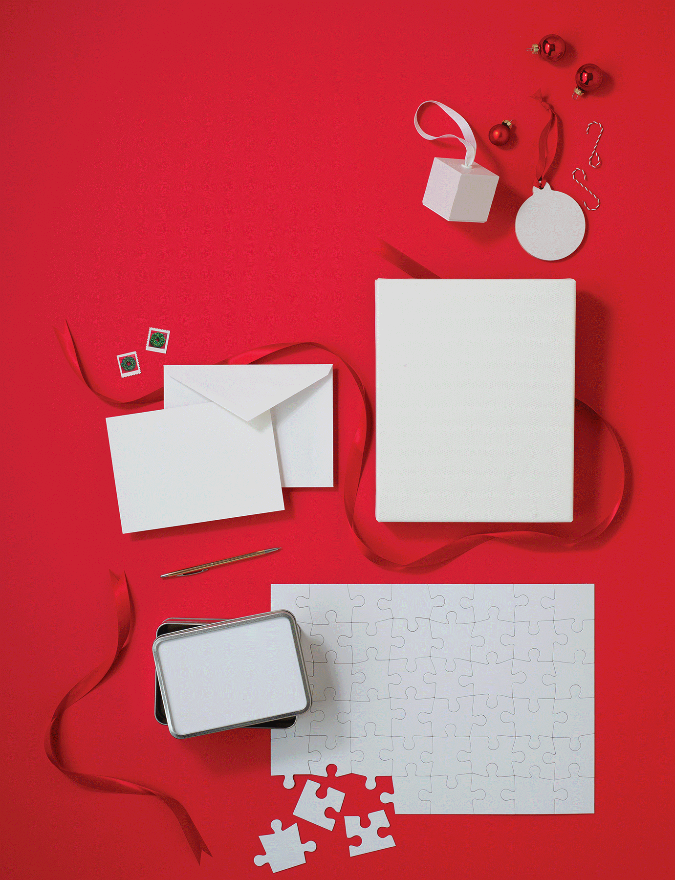 Flat lay of white Christmas-themed objects on a red background, including a white gift box with ribbon, small red ornaments, a white ornament with red ribbon, two small gift tags, an envelope, a pen, a puzzle with some pieces separated, and a container of blank game cards.