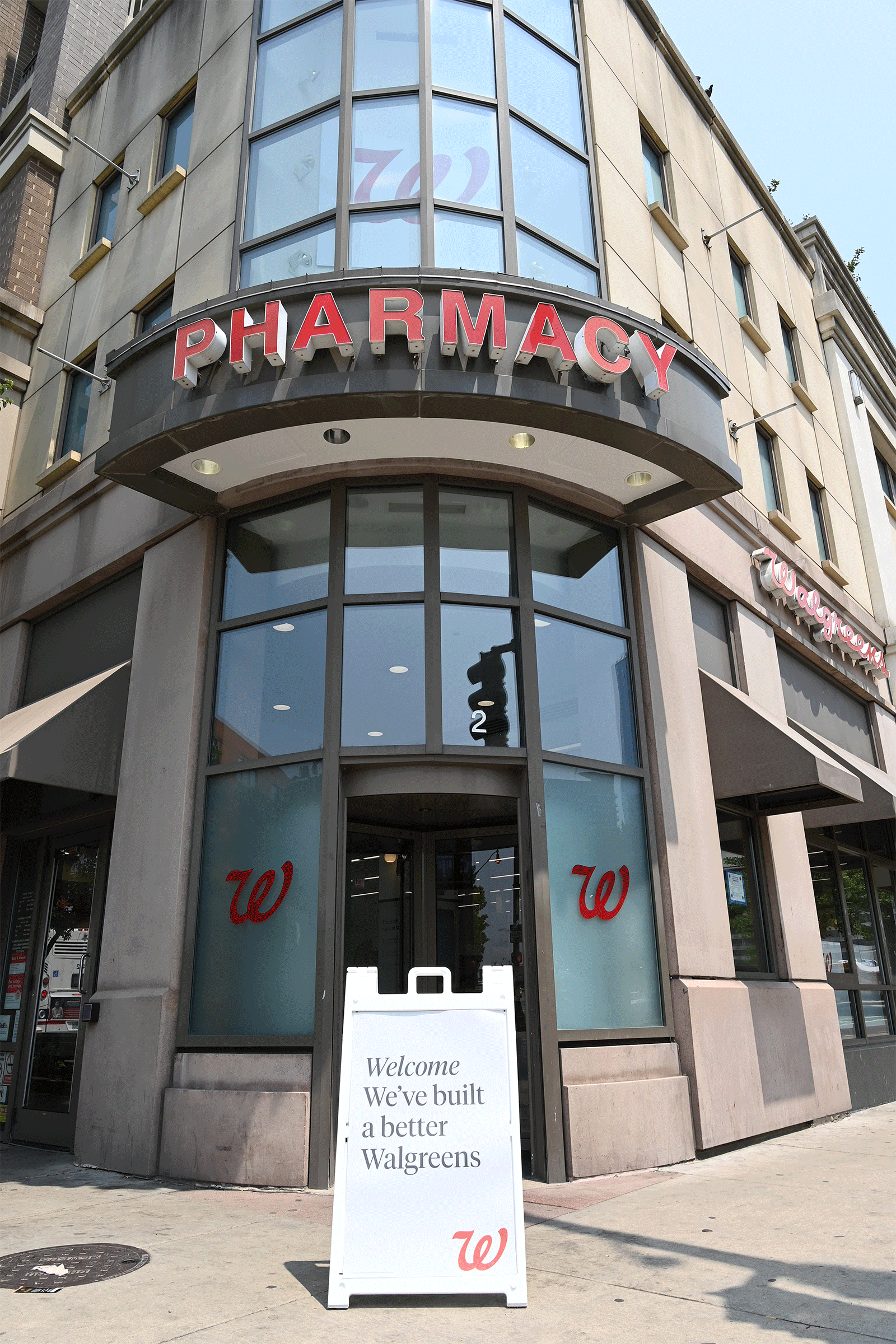 Outside Walgreens pharmacy with a large 'PHARMACY' sign above the entrance and a white signboard on the sidewalk that reads, 'Welcome We’ve built a better Walgreens'.