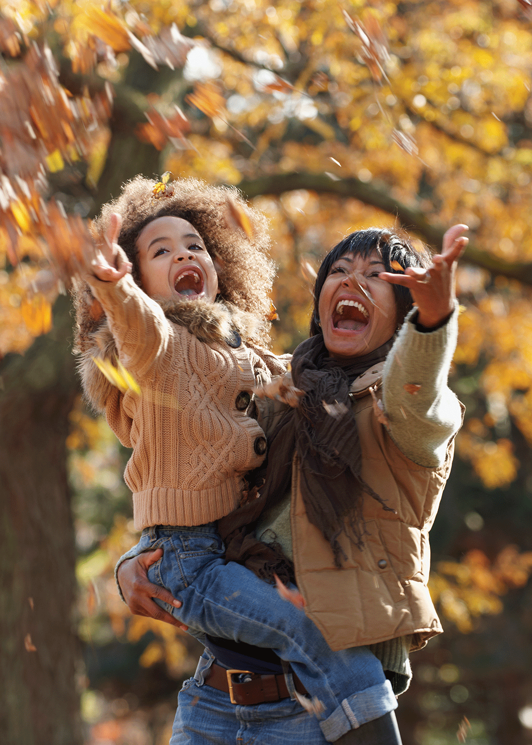 A woman holding a young girl outdoors playing in falling leaves on a sunny autumn day.