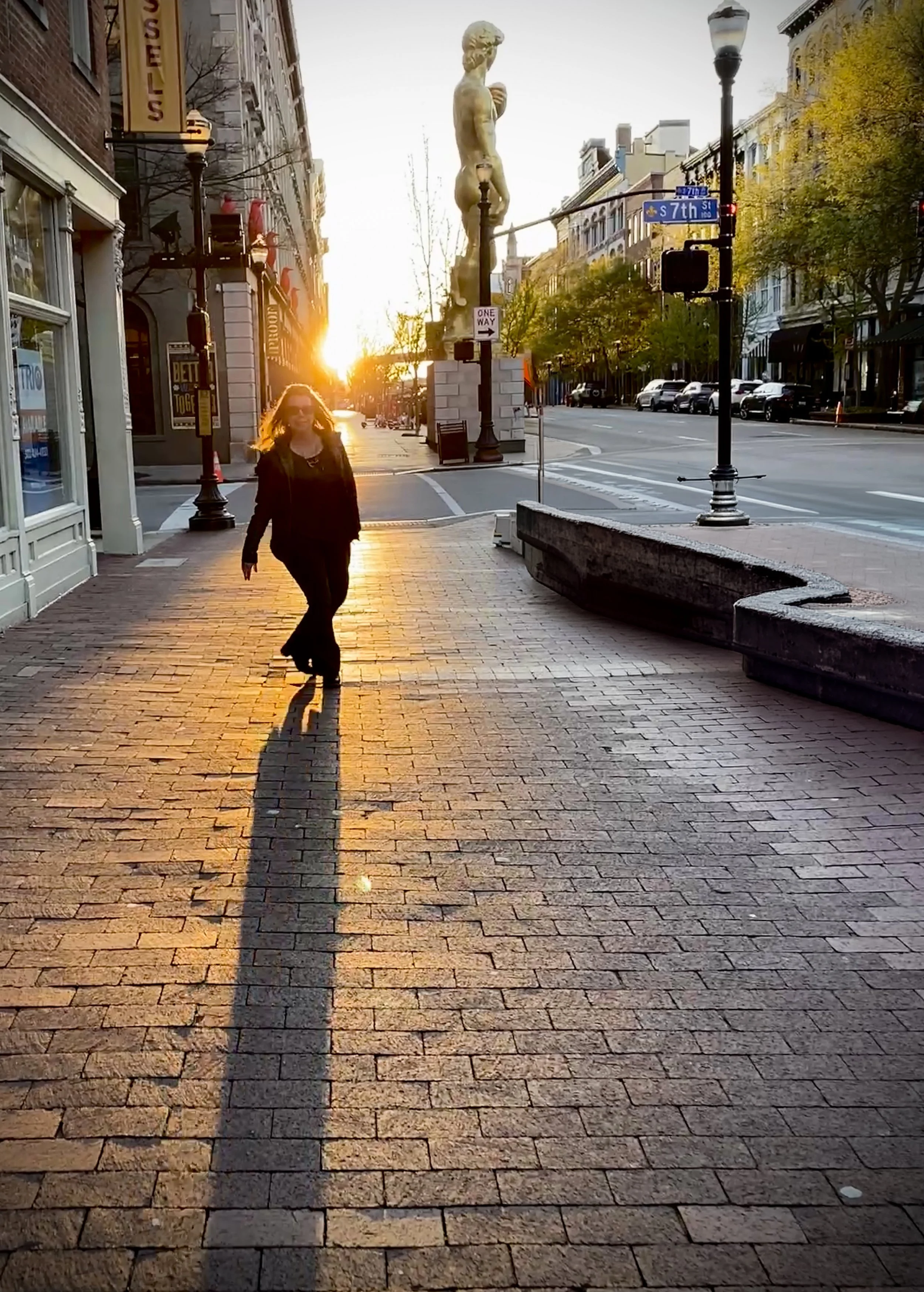 A woman walking on a brick sidewalk at sunset in a city with historic buildings, lampposts, and a large statue of a nude figure on a column in the background.
