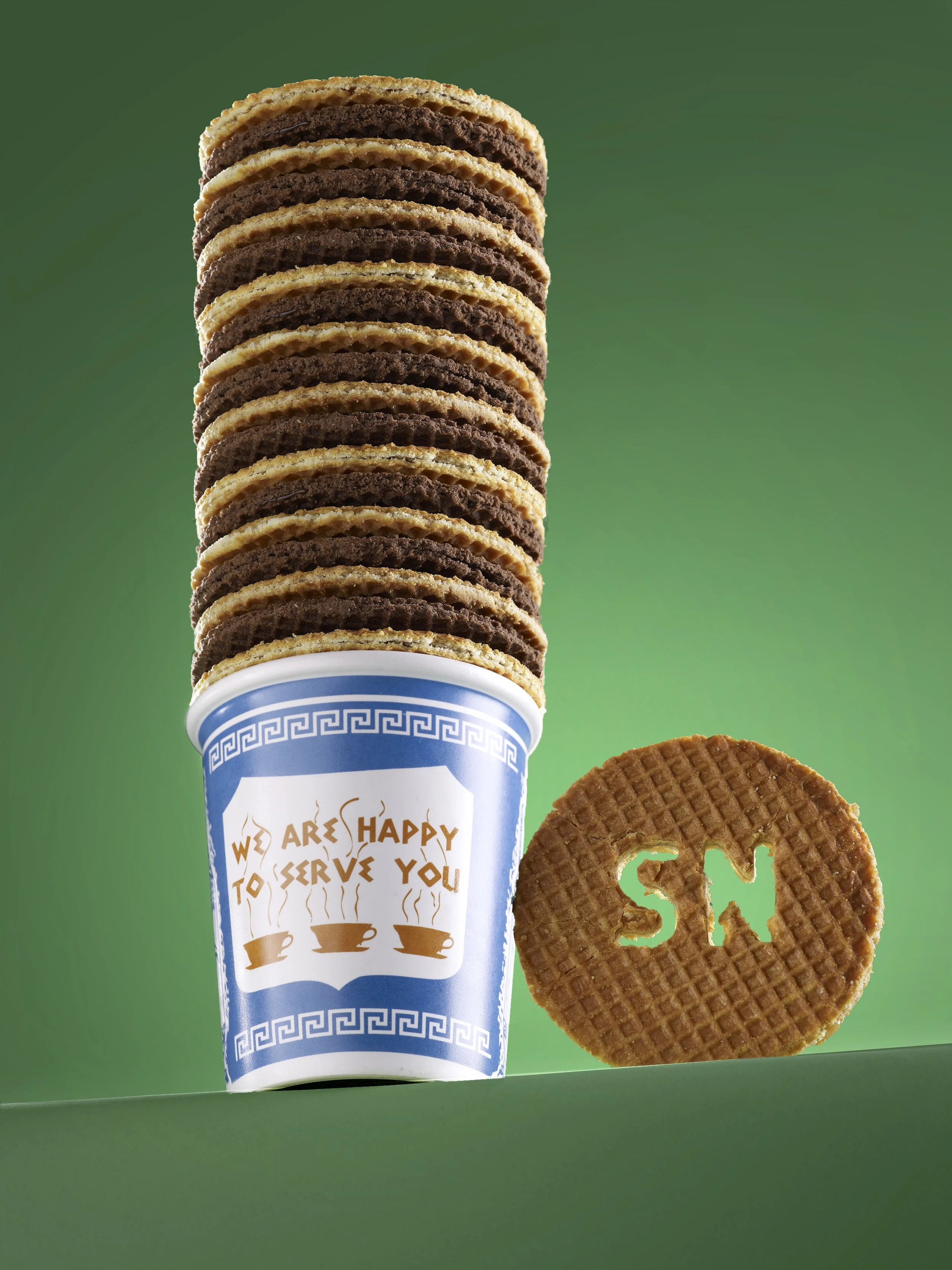 Stack of chocolate and vanilla cookies in a blue paper cup with a humorous message, next to a round caramel wafer cookie with the letter 'S' and 'N' cut out, against a green background.