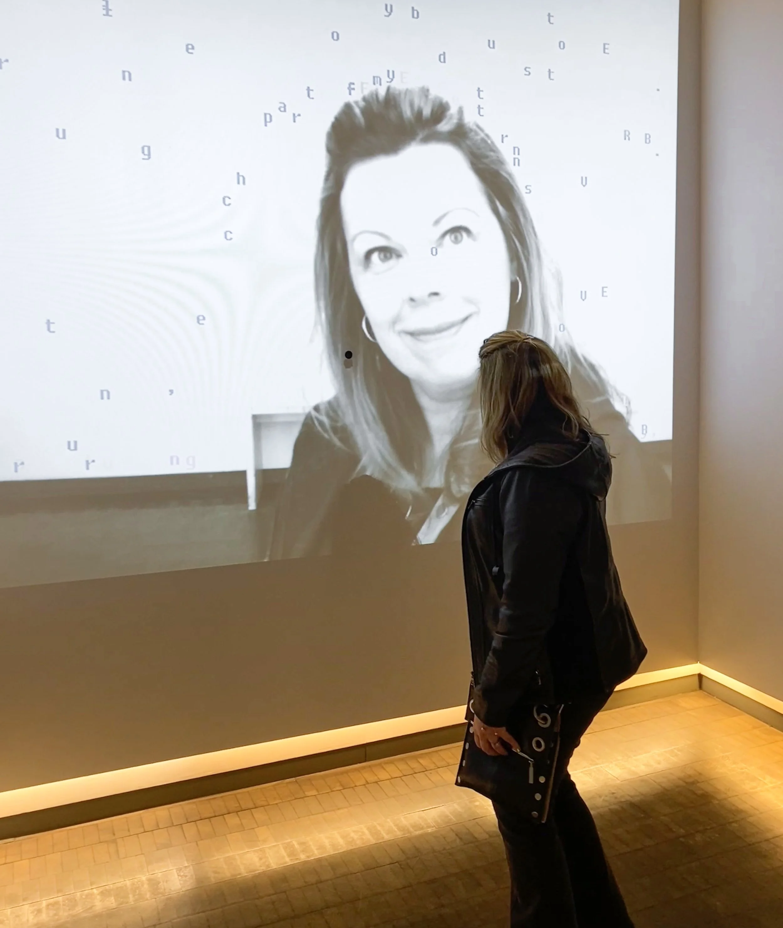 A woman in dark clothing standing in front of a large black and white digital portrait of a woman with text scattered around her face.