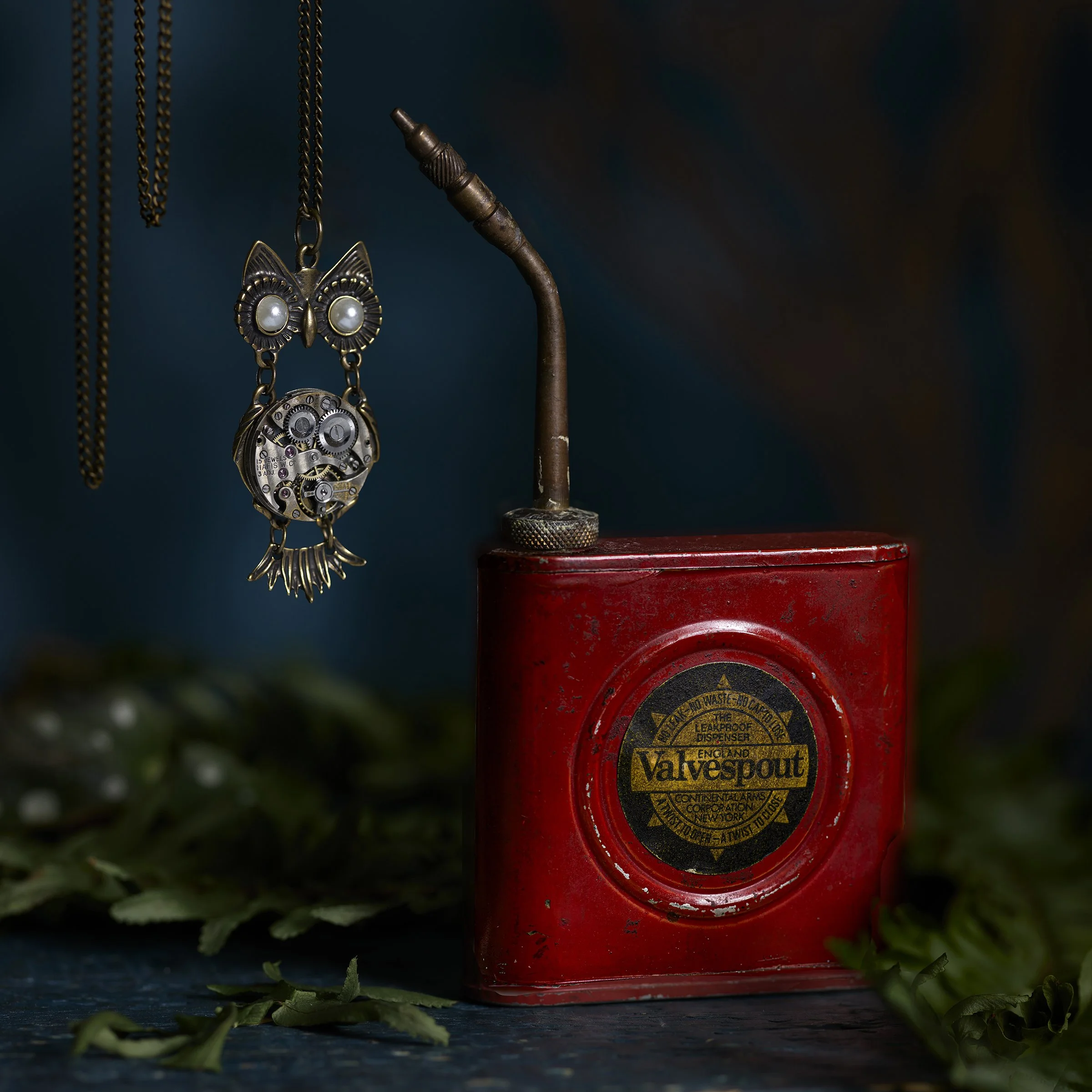 A vintage red Valvespout oil can with a metal spout, surrounded by green leaves. Hanging from a chain nearby is a decorative owl pendant with gears and pearls.
