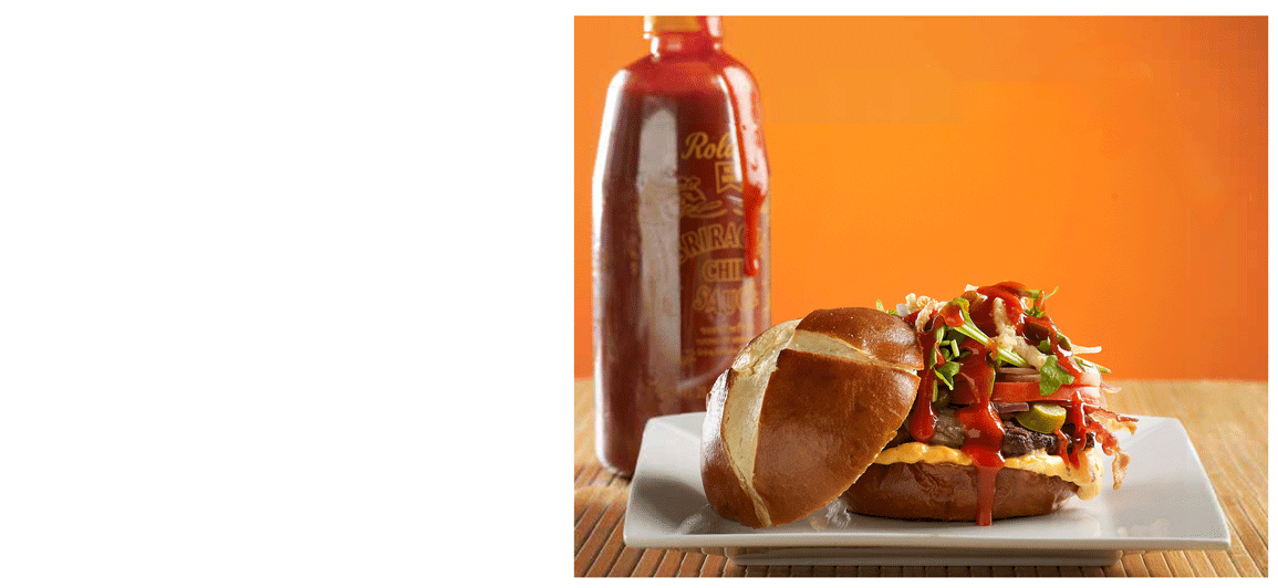 A burger on a white plate with lettuce, tomato, cheese, bacon, and ketchup, served with a bottle of hot sauce in the background on an orange wall.