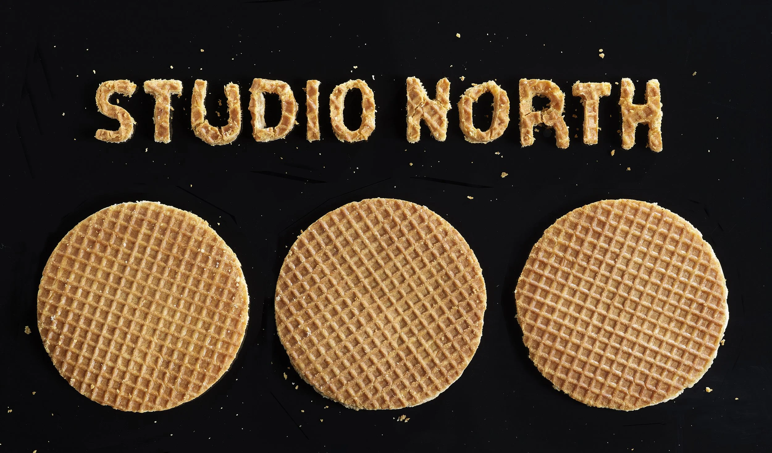 The words 'STUDIO NORTH' are spelled out in cookie crumbs over three round cookies with a waffle pattern on a black background.