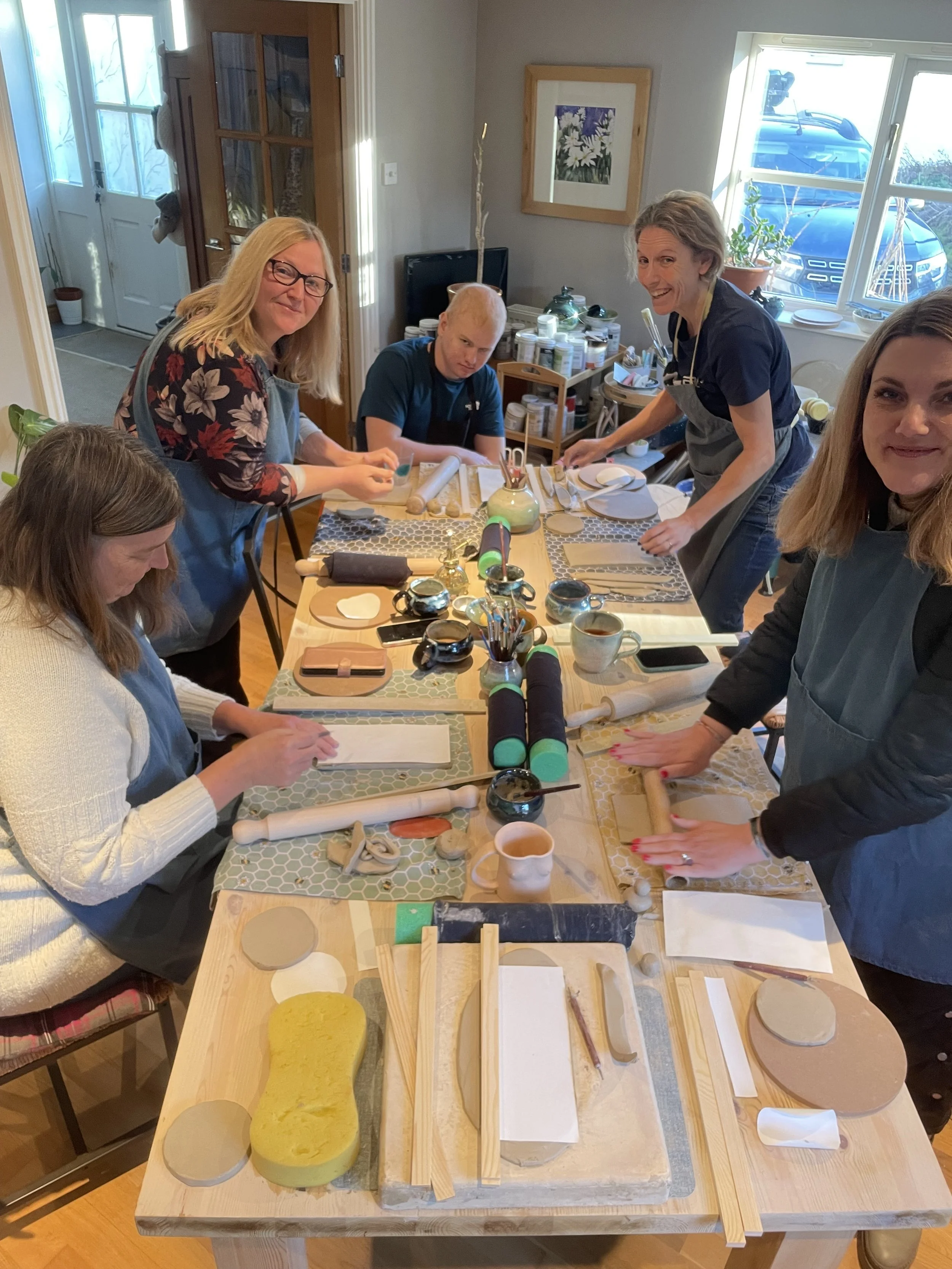 Pottery Experience.  Pottery Fun.  Pottery Class.  Pottery Mug Making. Pottery lessons. Group of women and a young man gathered around a table with clay and pottery tools, preparing for a pottery or ceramics activity in a cozy indoor setting.