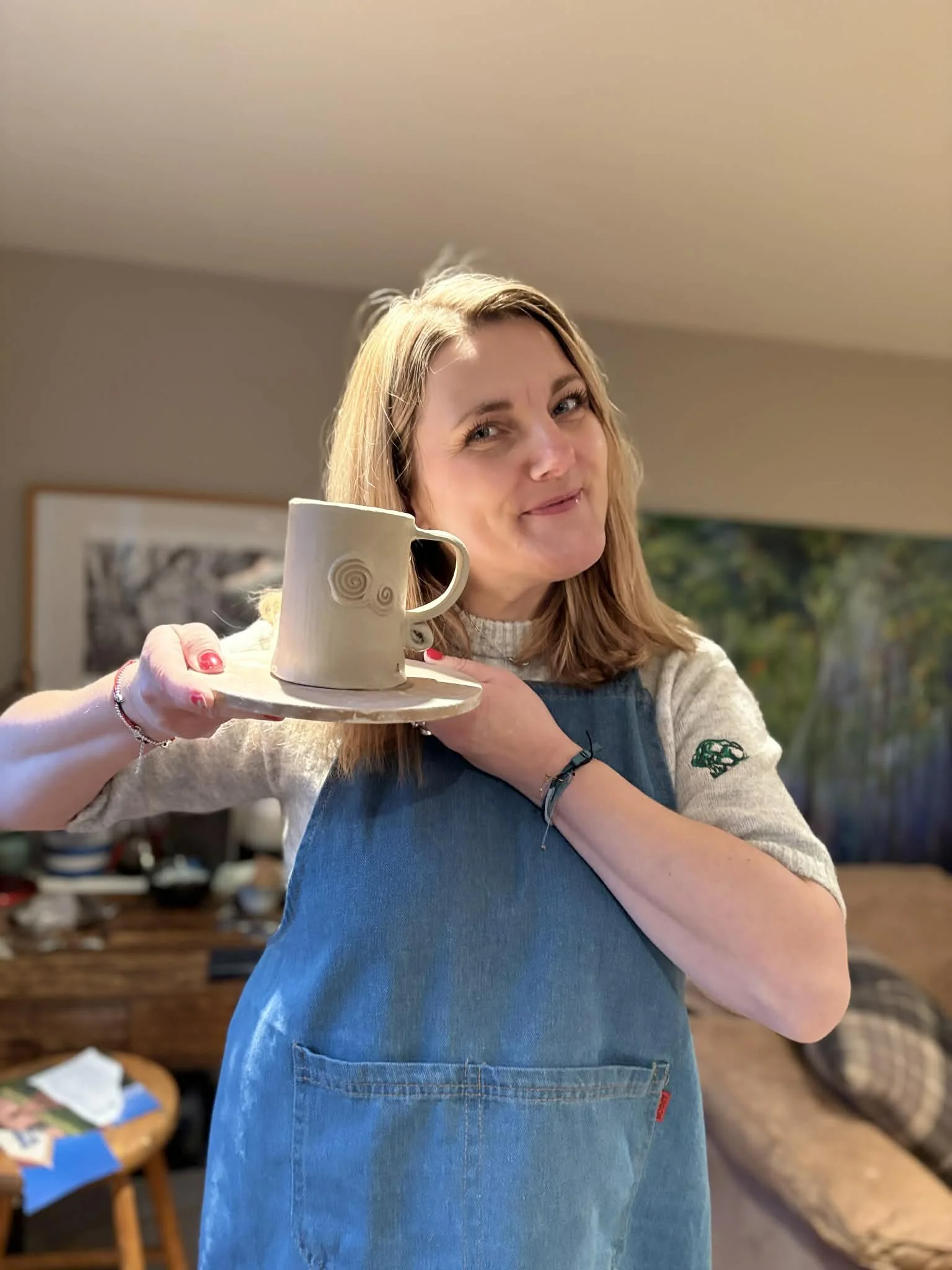 Happy Pottery Woman with shoulder-length blonde hair smiling, holding a ceramic mug on a saucer, wearing a gray shirt and a blue denim apron in a cozy room. Pottery class.  Learn Pottery. Pottery fun. Mug Making. Pottery Experience. Pottery Lessons.