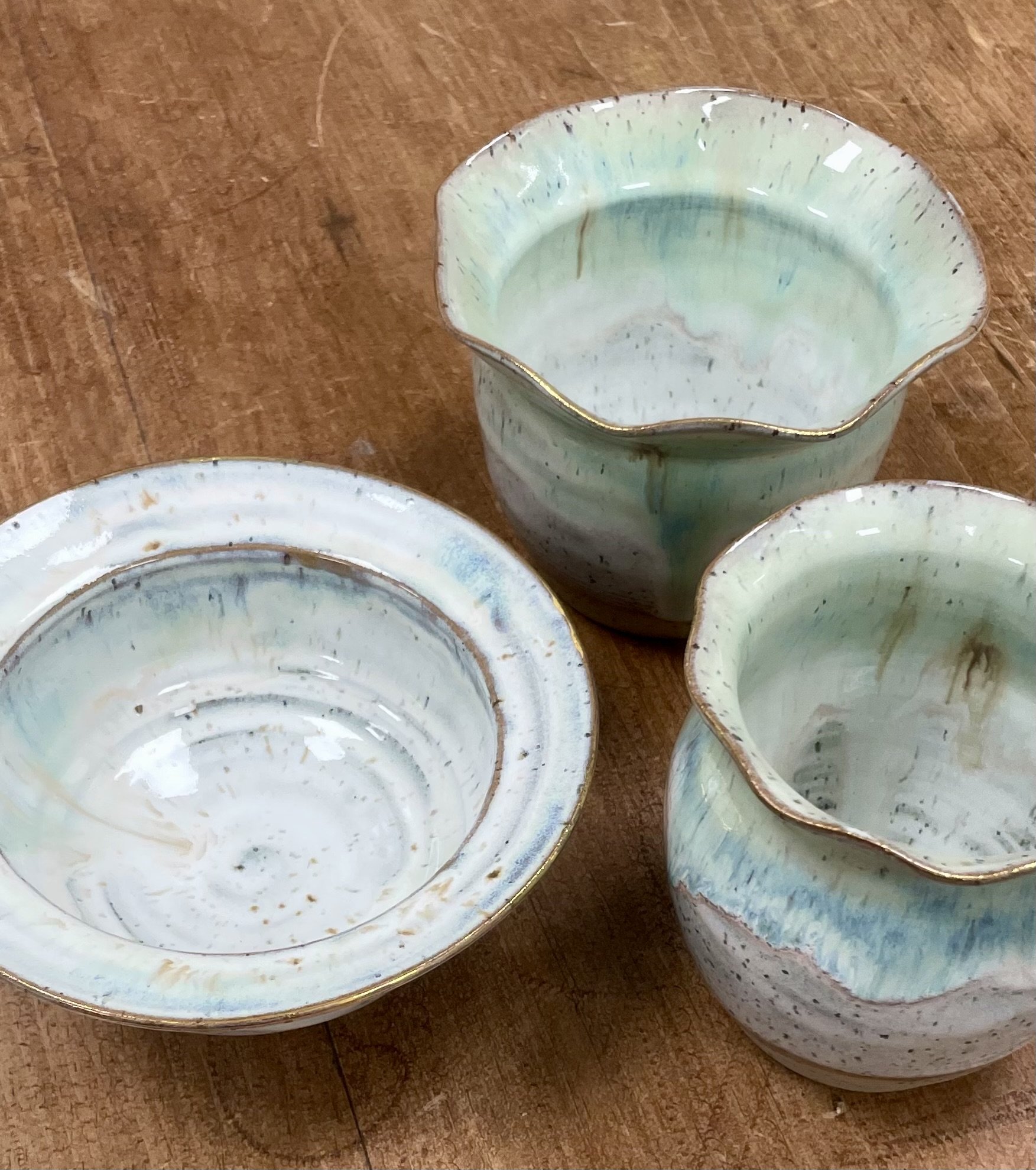 Set of three handmade ceramic bowls with a blue and green glaze, placed on a wooden surface, with one bowl upside down showing the interior and slight gold detailing along the rims.