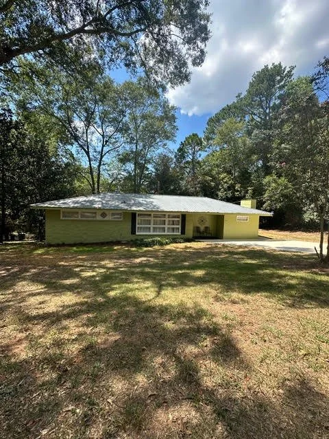 A single-story house painted light green with a metal roof, surrounded by trees and a grassy yard.