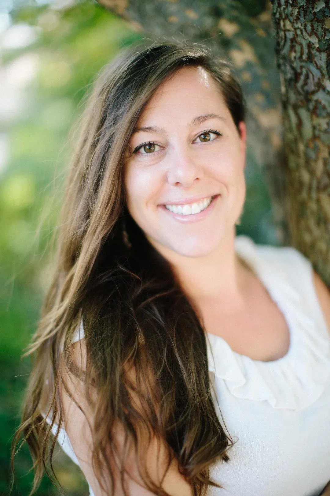 A woman with long brown hair smiling, leaning against a tree outdoors with a blurred green background.