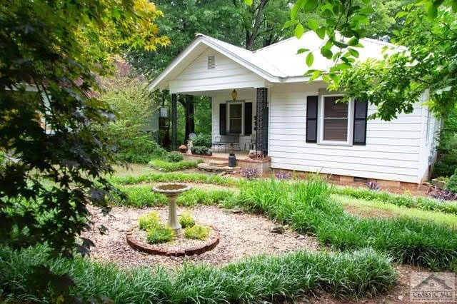 White house with black shutters and a small front porch surrounded by green trees and plants, with a decorative birdbath in the front yard.