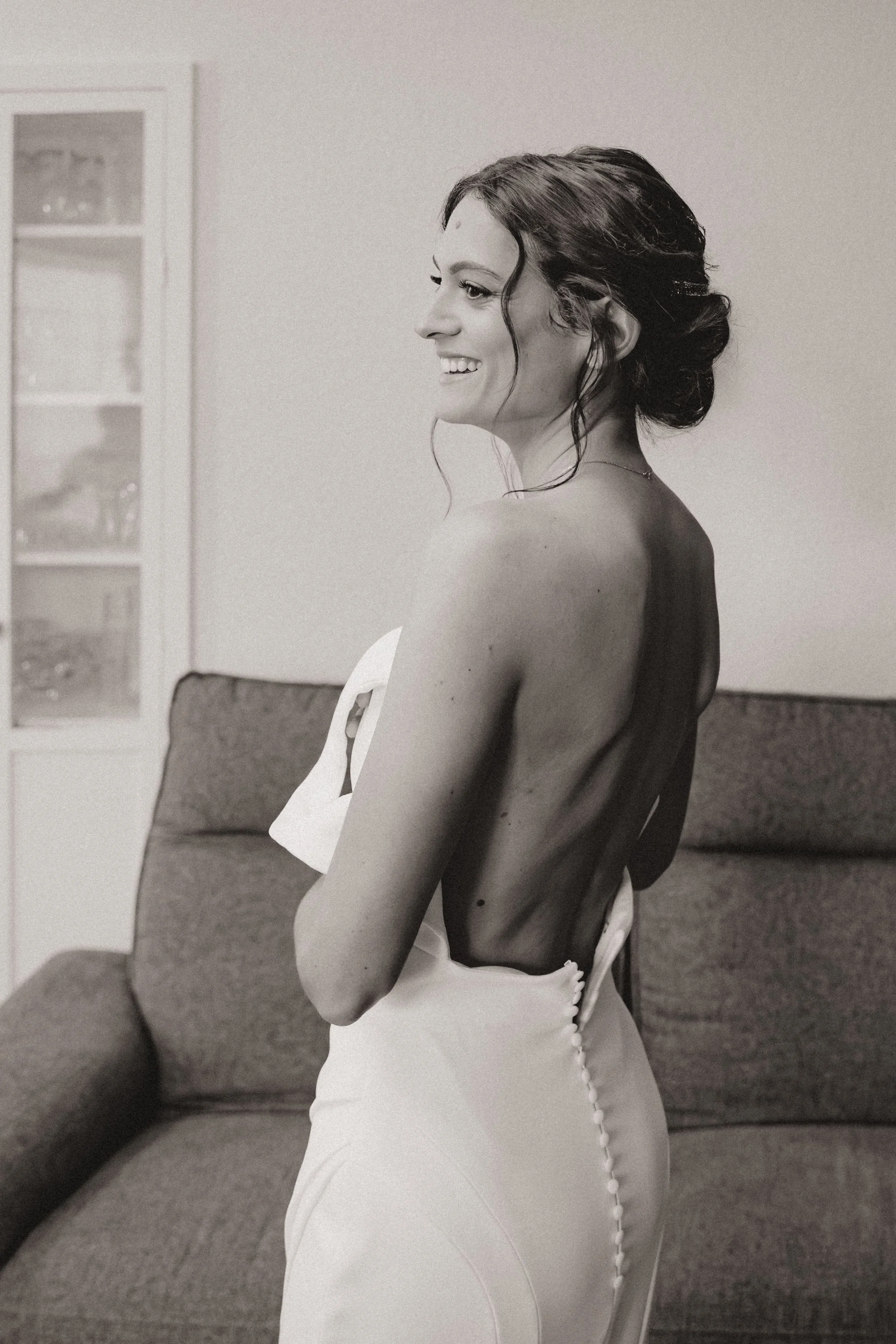 Une femme souriante, vêtue d'une robe blanche, avec les épaules découvertes, se tient à côté d'un canapé dans une pièce intérieure. Elle porte un maquillage naturel et élégant pour son mariage.