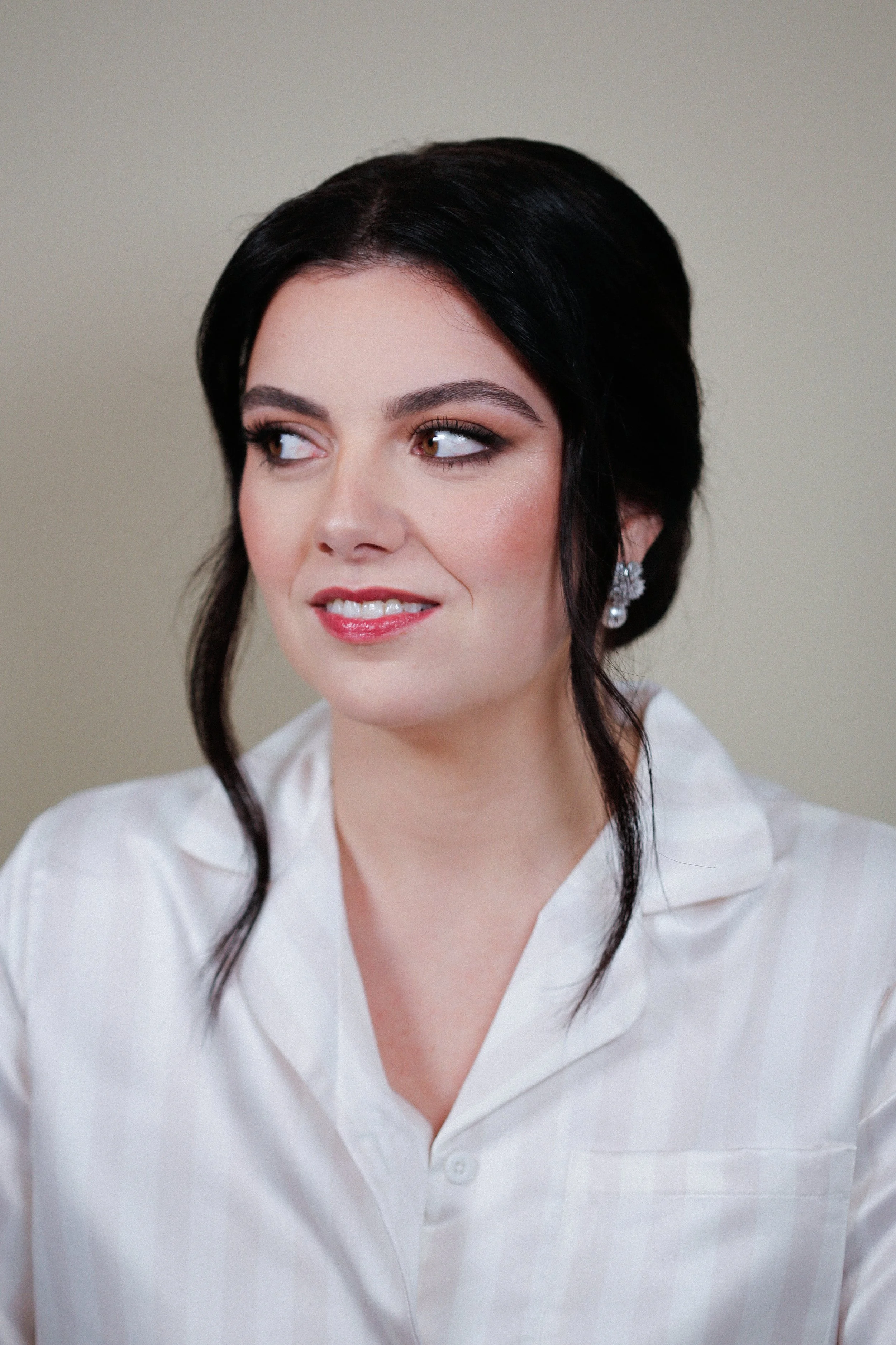 Une femme avec des cheveux noirs et une chemise blanche, souriant légèrement, regardant de côté. Elle porte un maquillage intense , glamour et élégant pour son mariage d'hiver.