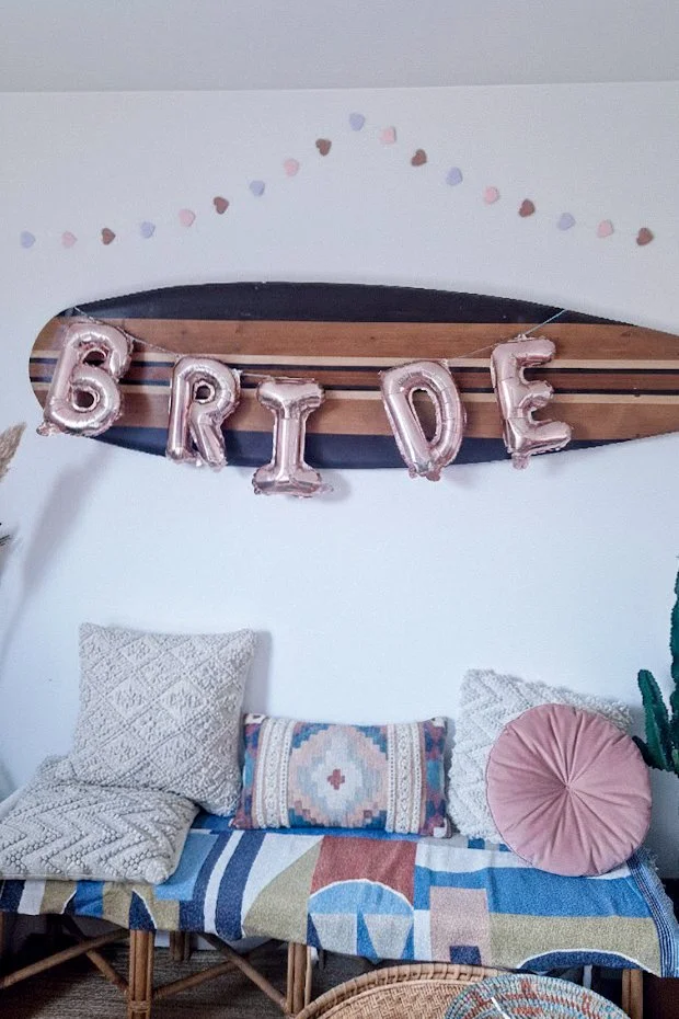 Décoration de fête avec des ballons en forme de lettres formant le mot 'BRIDE', suspendus devant une planche en bois décorée dans un salon avec canapé et coussins.