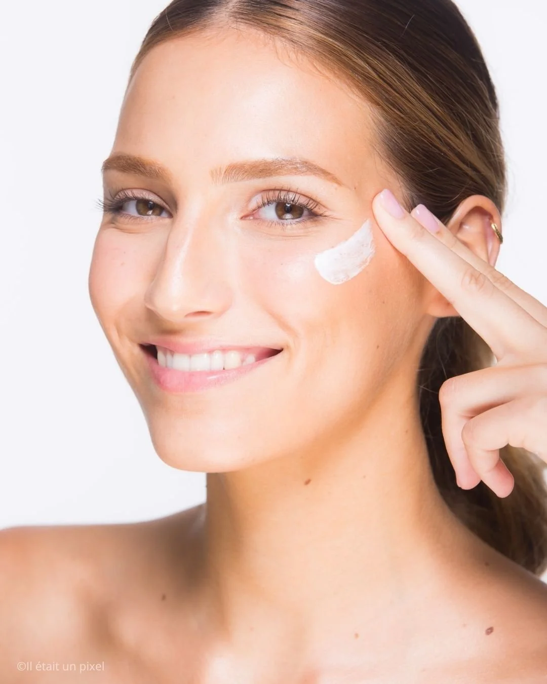 Une femme souriante appliquant une crème ou un produit de soin sur sa joue droite avec son index, face à un fond blanc.