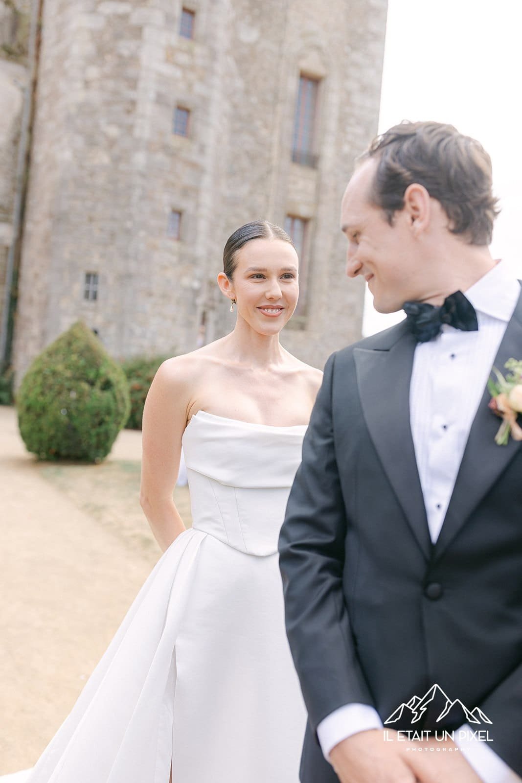 Un couple marié lors d'une séance photo à l'extérieur, avec un château en arrière-plan. La femme porte une robe de mariée blanche et le homme un costume noir avec nœud papillon. Elle porte un maquillage naturel et élégant pour son mariage.