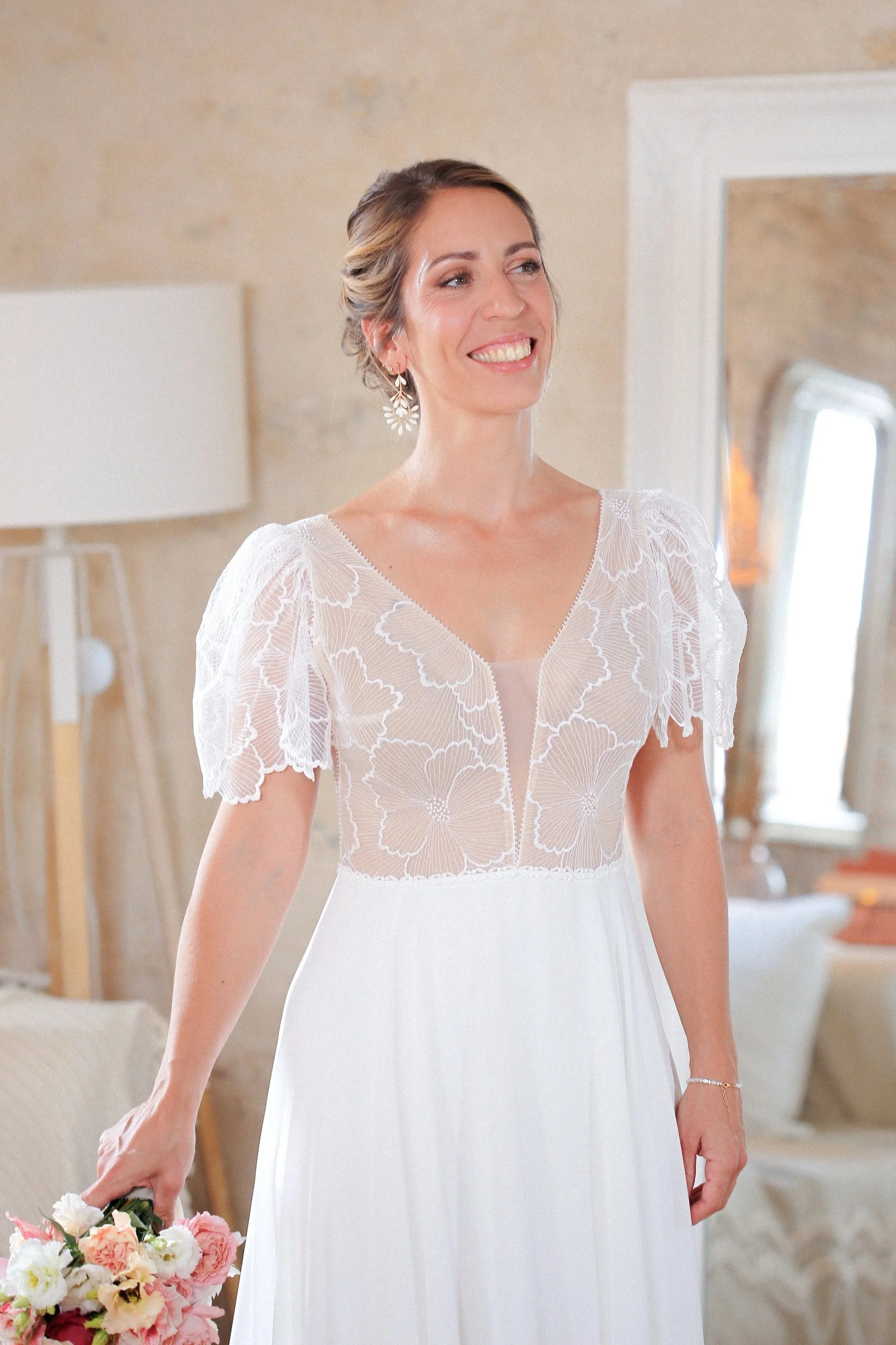 Femme souriante en robe de mariée blanche avec dentelle, tenant un bouquet de fleurs roses et blanches, dans une pièce lumineuse. Elle porte un maquillage naturel et élégant pour son mariage.