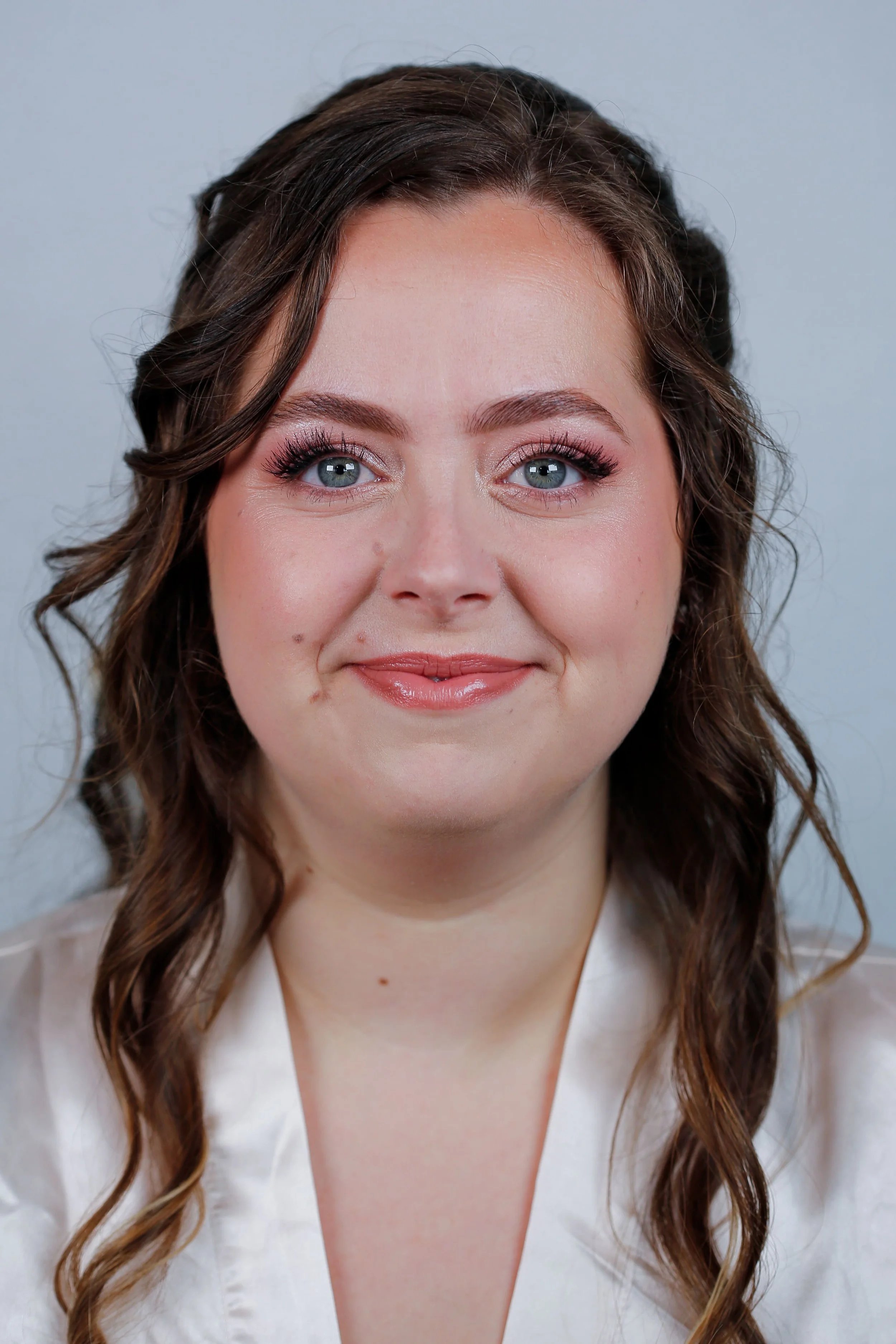 Portrait d'une femme souriante avec des cheveux bruns bouclés et des yeux bleus, portant un chemisier blanc, contre un fond gris clair. Elle porte un maquillage naturel et frais pour son mariage.