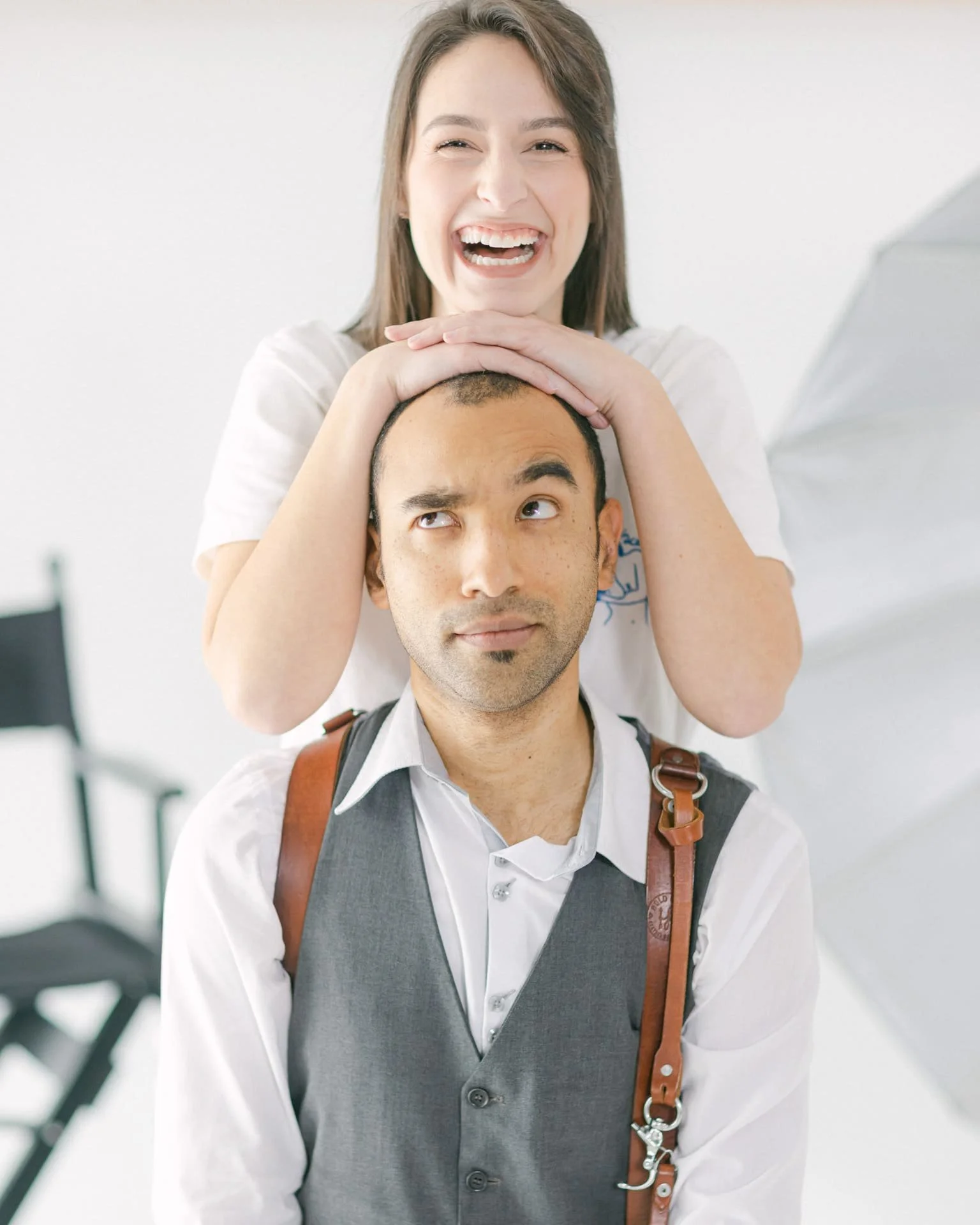 Une femme souriante pose ses mains sur la tête d'un homme qui regarde de côté, dans une ambiance lumineuse de studio . Elle est maquilleuse et lui photographe.