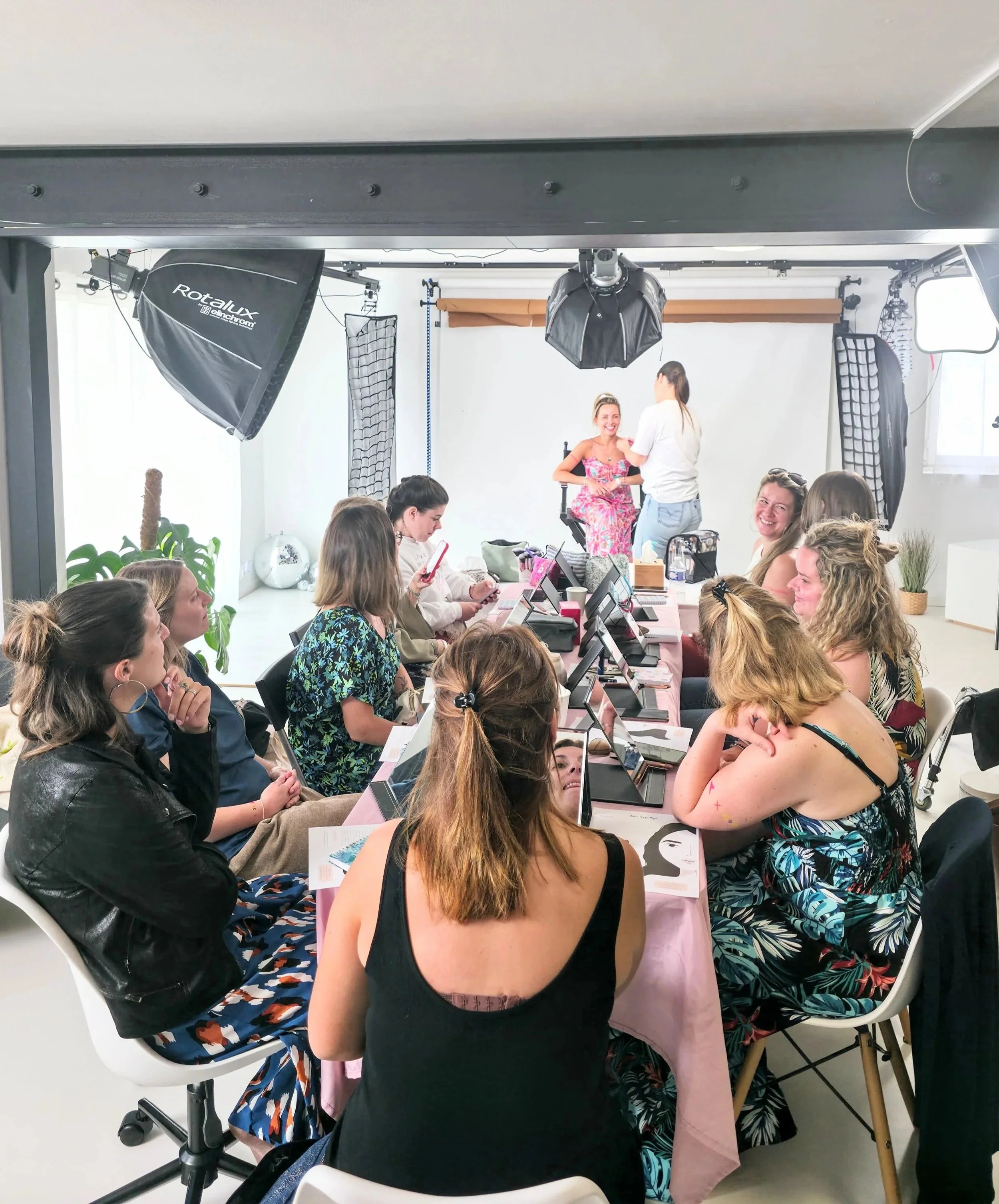 Un atelier de maquillage pour un evjf dans un studio. un groupe de femmes assises autour d'une table, tandis qu'une femme en robe rose se fait maquiller et une autre explique les techniques de maquillage, avec un éclairage professionnel.