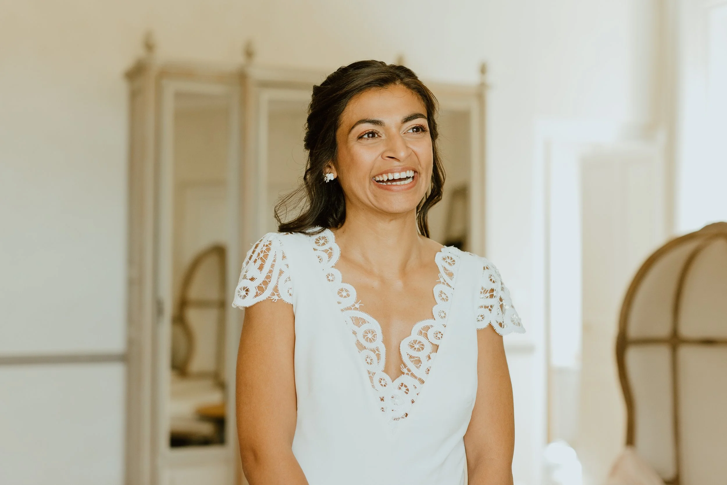 Femme souriante portant une robe blanche avec des détails en dentelle, dans une pièce lumineuse.
