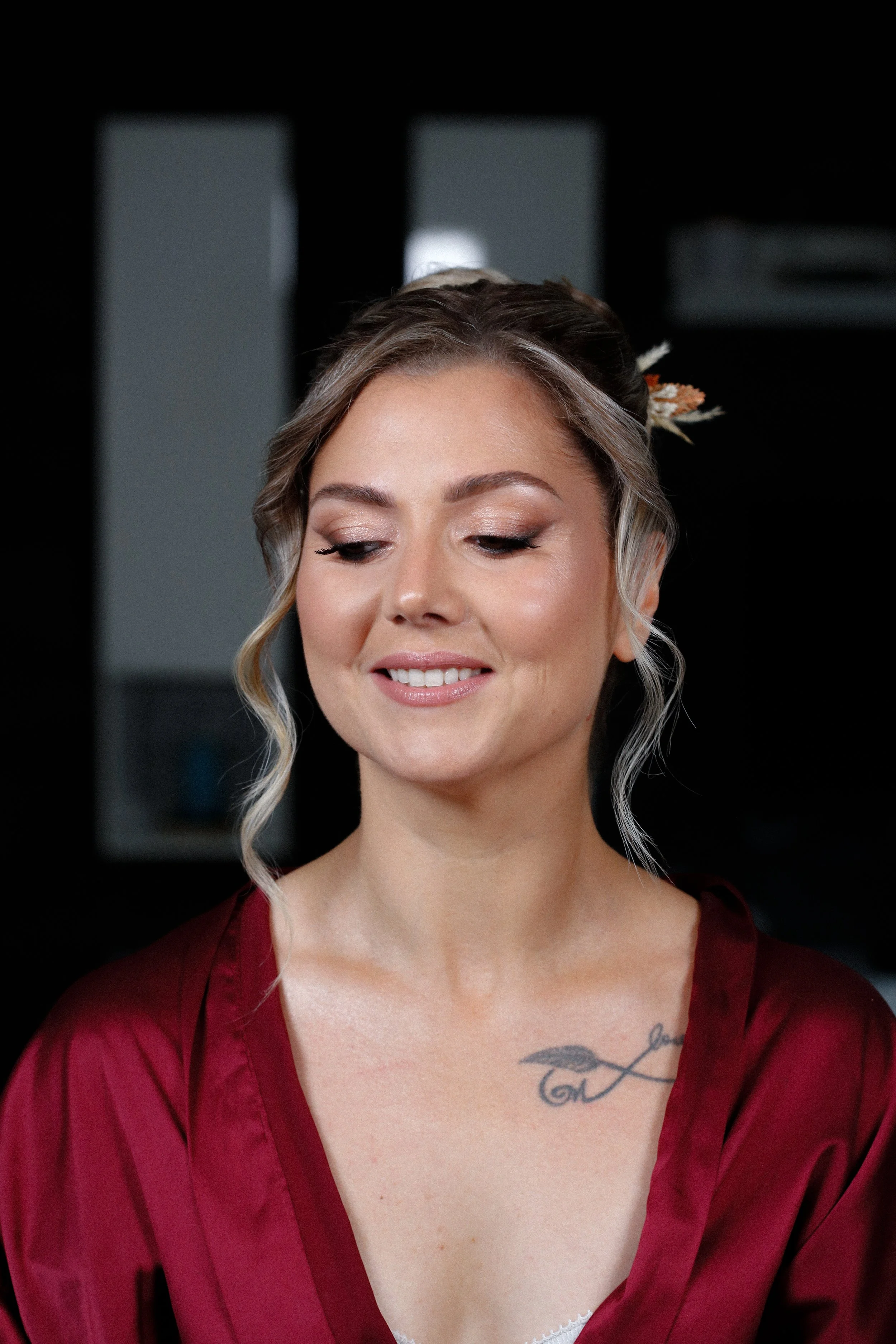 Une femme avec des cheveux coiffés, portant une robe rouge en satin, souriante, avec un tatouage d'une plume sur la poitrine, dans un intérieur sombre.