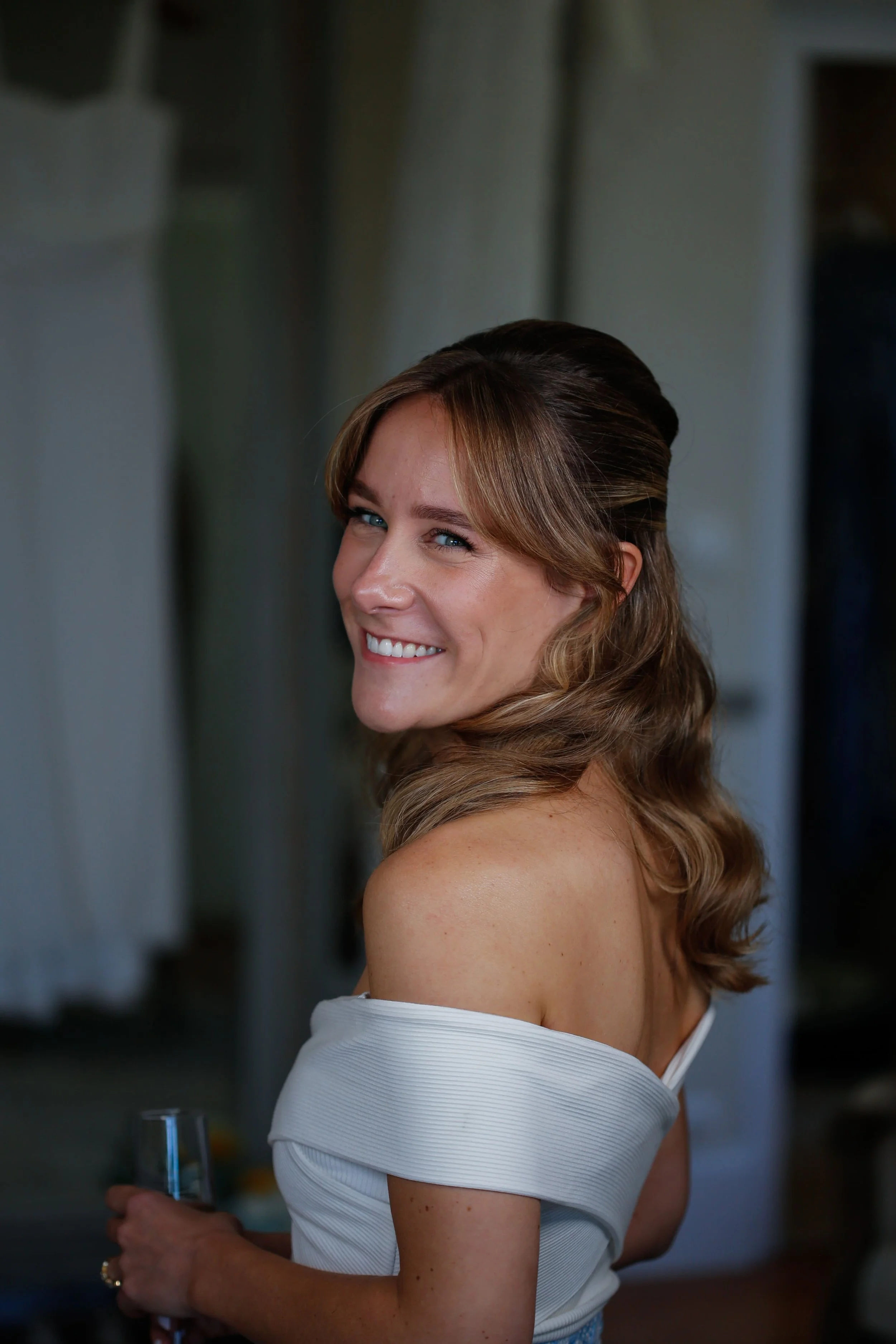 Femme souriante avec cheveux ondulés, portant une robe blanche, tenant un verre à la main, dans un intérieur.