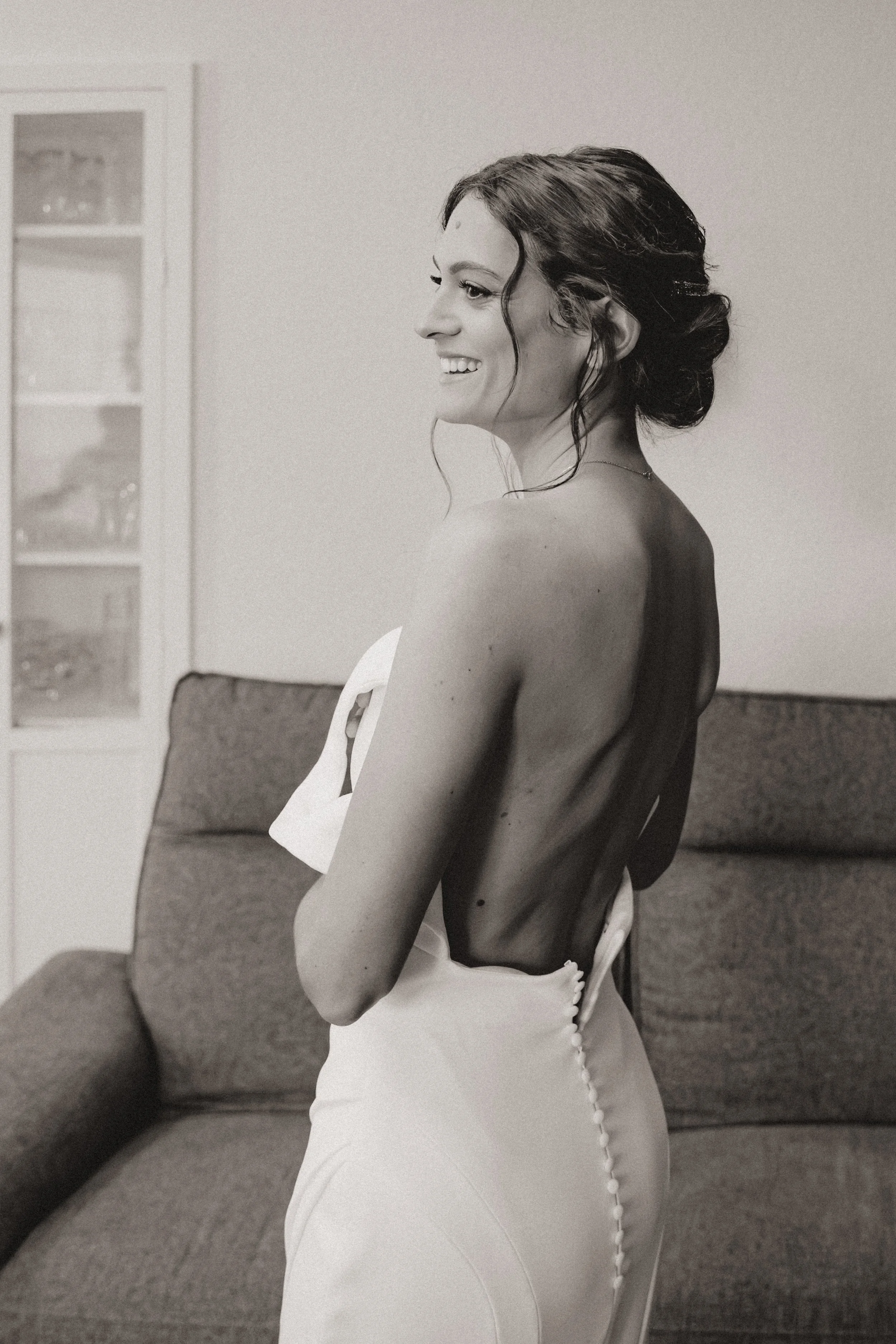 Une femme souriante, vêtue d'une robe blanche, avec les épaules découvertes, se tient à côté d'un canapé dans une pièce intérieure. Elle porte un maquillage naturel et élégant pour son mariage.