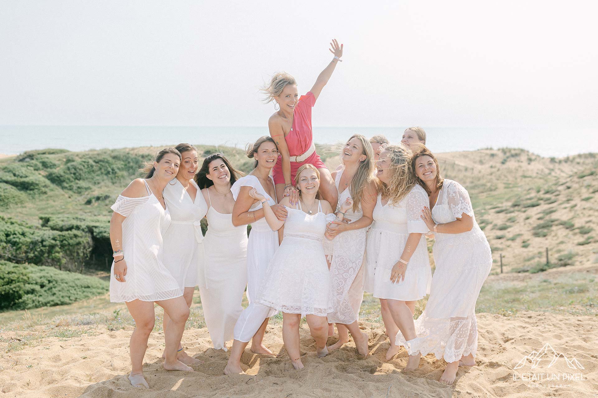 Un groupe de femmes en robes blanches, certaines avec des détails en crochet, pose sur la plage. Une femme en robe rose est assise sur plusieurs autres femmes, levant un bras en l'air, toutes souriantes dans un décor en plein air avec du sable et des collines verdoyantes en arrière-plan.