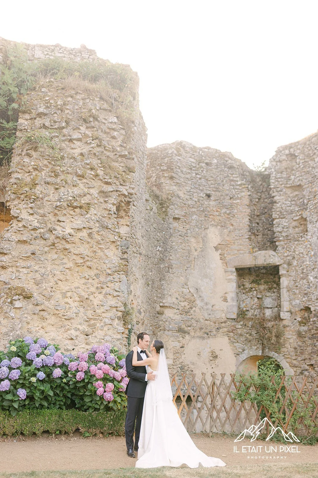 Un couple de mariés, un homme en costume noir et une femme en robe de mariage blanche, se tiennent dans un éclat de pierre ancienne, entourés de fleurs de hortensia violettes, lors d'une séance photo de mariage en extérieur.