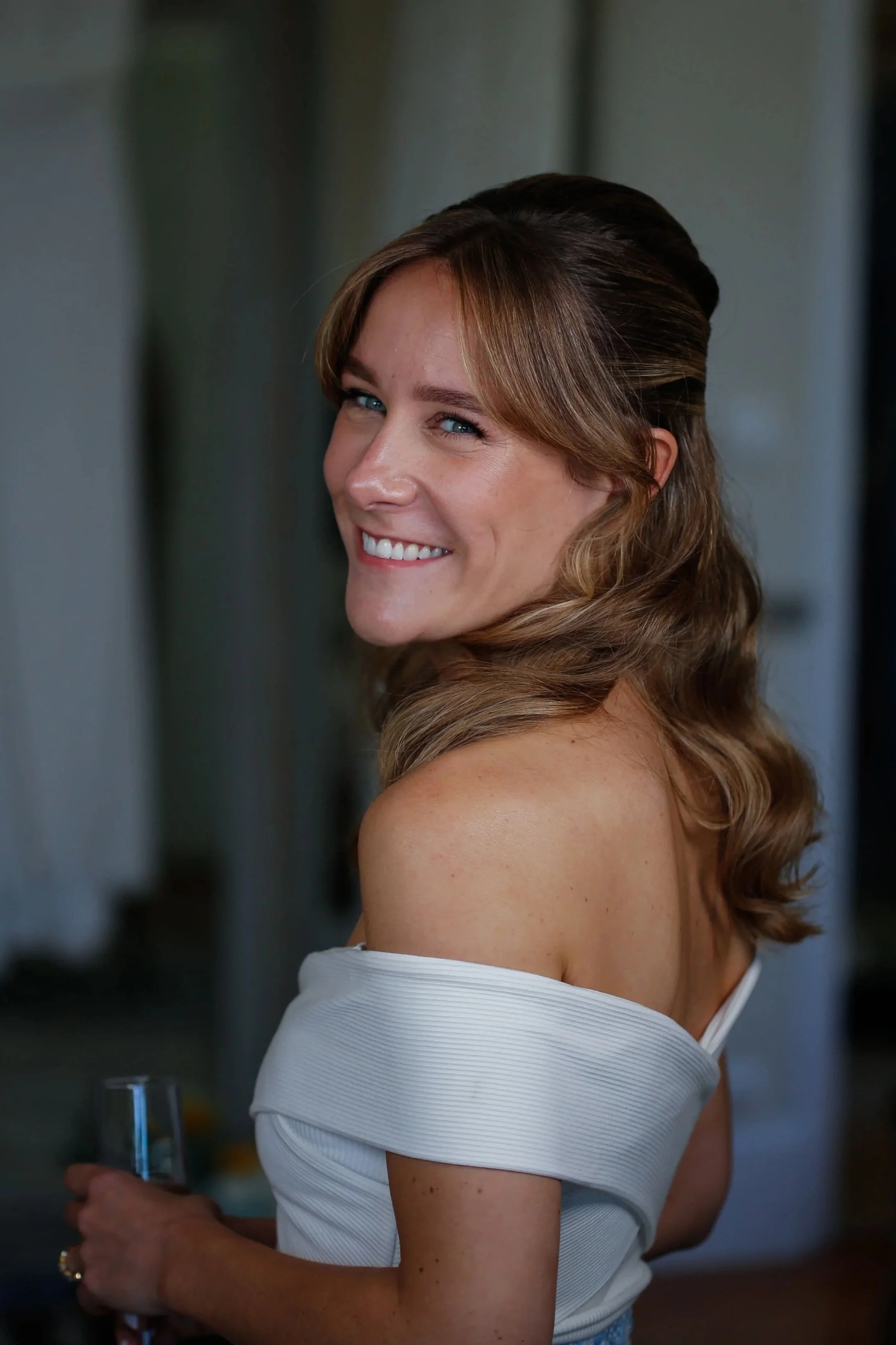 Femme souriante avec cheveux ondulés, portant une robe blanche, tenant un verre à la main, dans un intérieur.