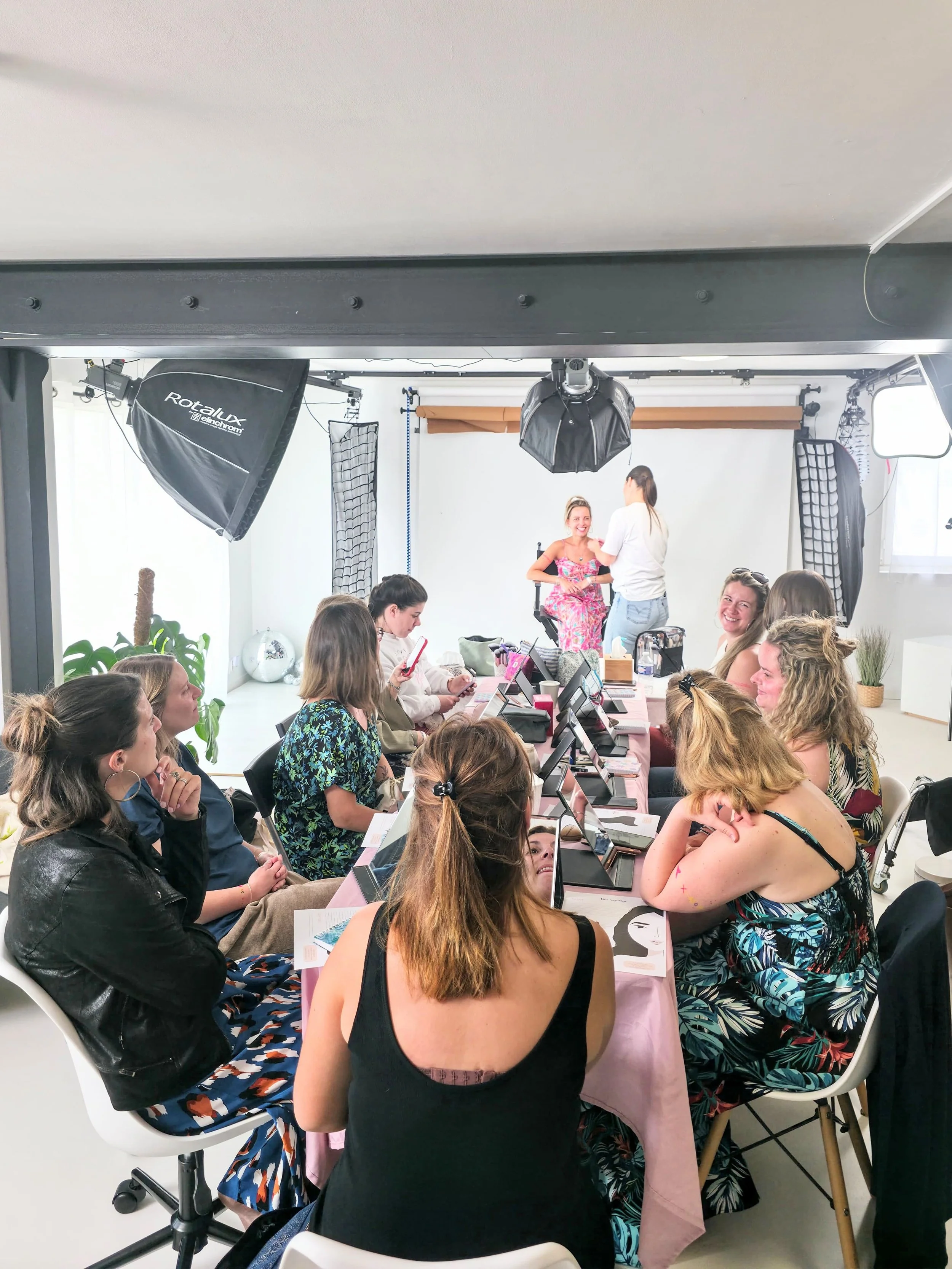 Un atelier de maquillage pour un evjf dans un studio. un groupe de femmes assises autour d'une table, tandis qu'une femme en robe rose se fait maquiller et une autre explique les techniques de maquillage, avec un éclairage professionnel.