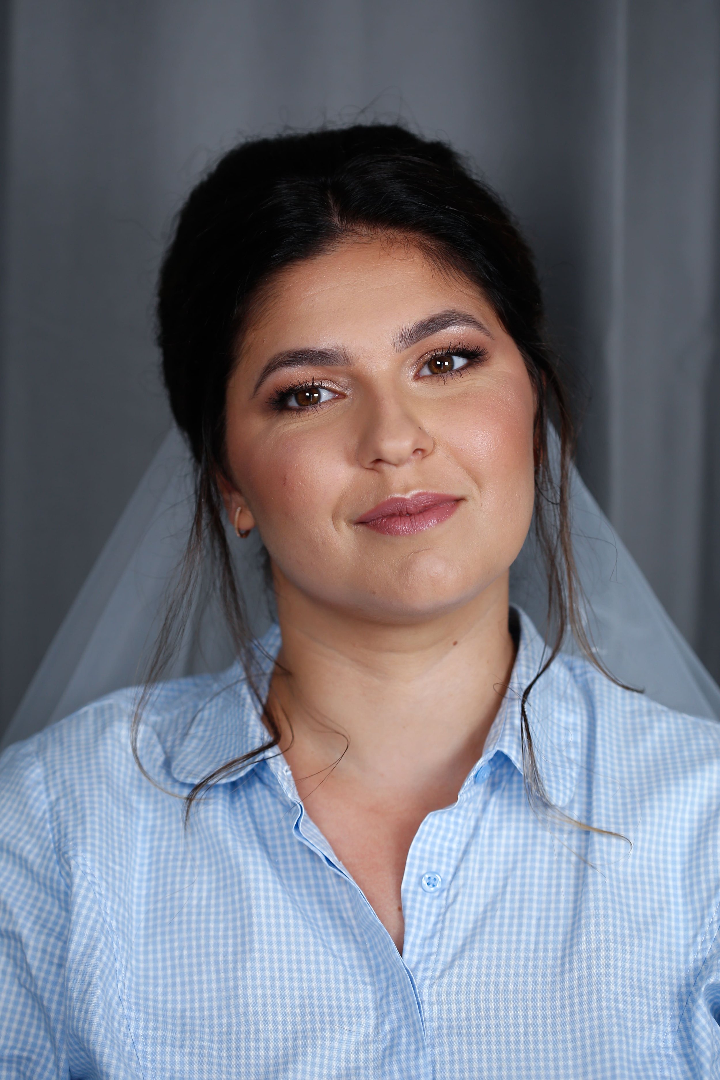 Une femme souriante porte une chemise à carreaux bleue et une voile blanche, avec un fond gris.