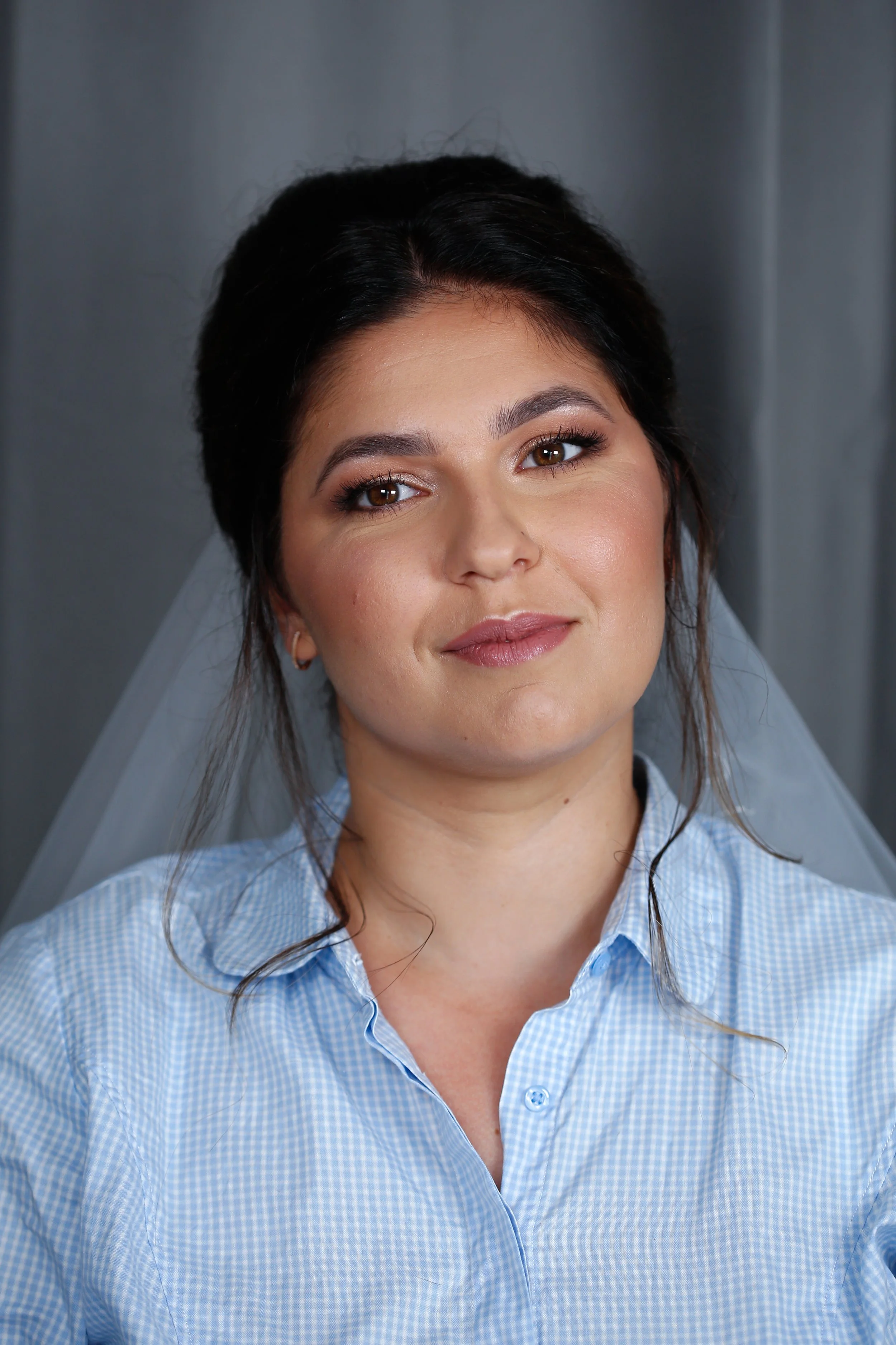 Une femme souriante porte une chemise à carreaux bleue et une voile blanche, avec un fond gris.