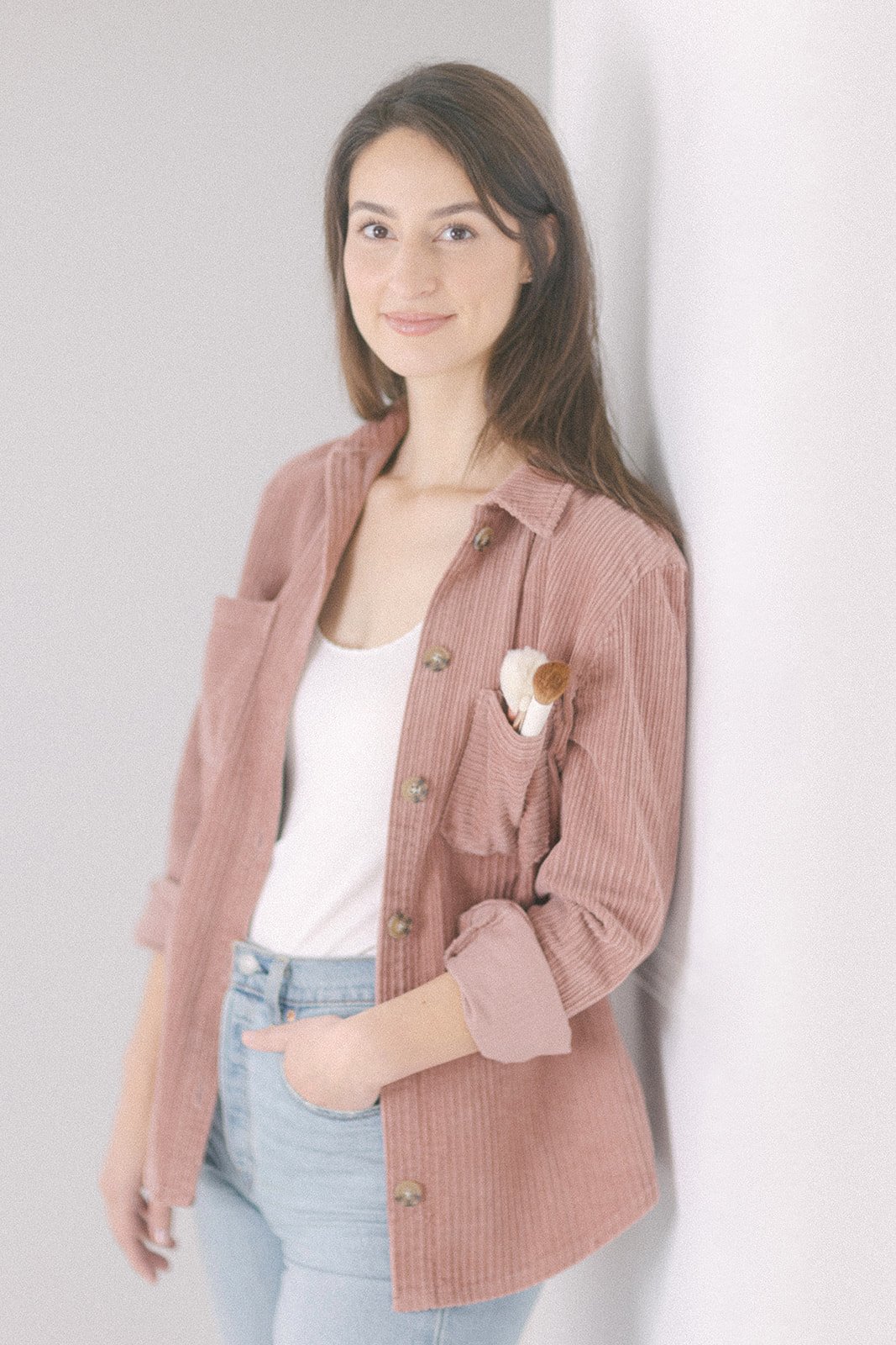 Jeune femme souriante en chemise rose en velours côtelé, avec pinceaux de maquillage dans la poche, posant contre un mur blanc. Elle est maquilleuse professionnelle.