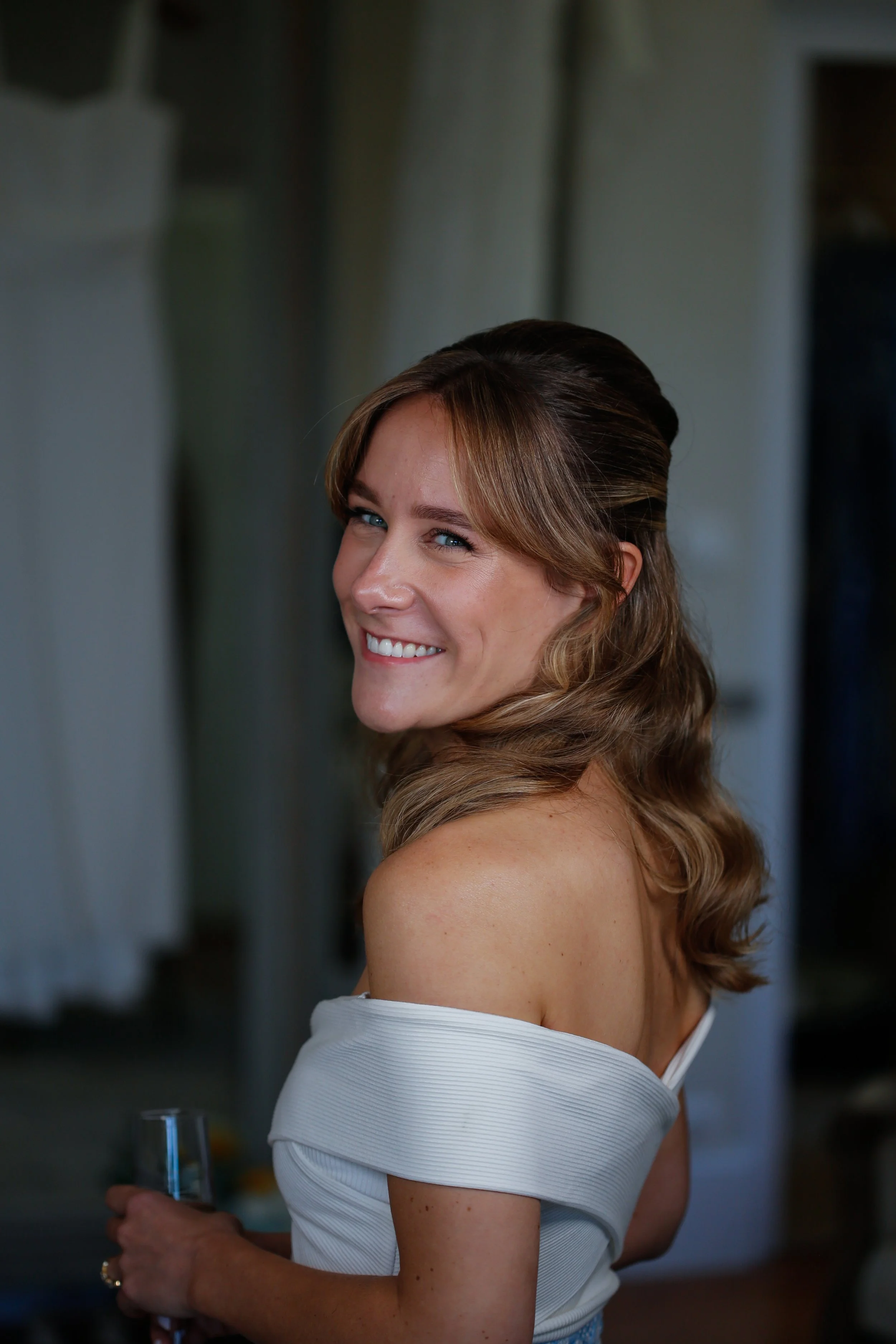 Femme souriante avec cheveux ondulés, portant une robe blanche, tenant un verre à la main, dans un intérieur.