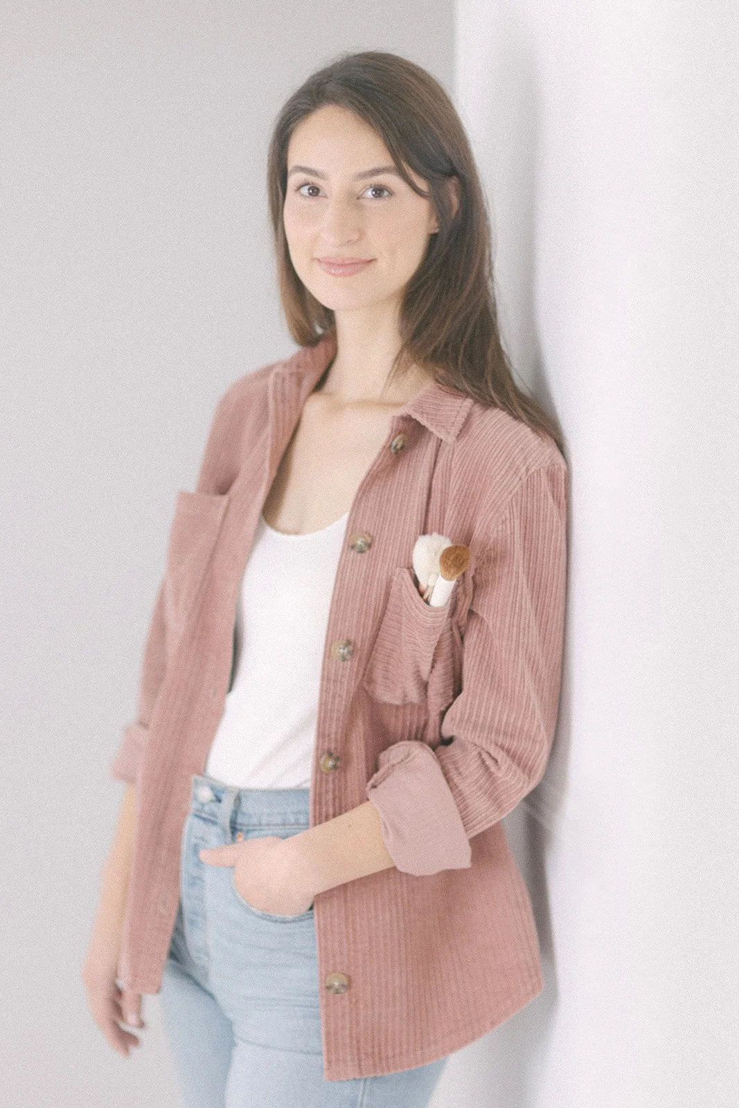 Jeune femme souriante en chemise rose en velours côtelé, avec pinceaux de maquillage dans la poche, posant contre un mur blanc. Elle est maquilleuse professionnelle.