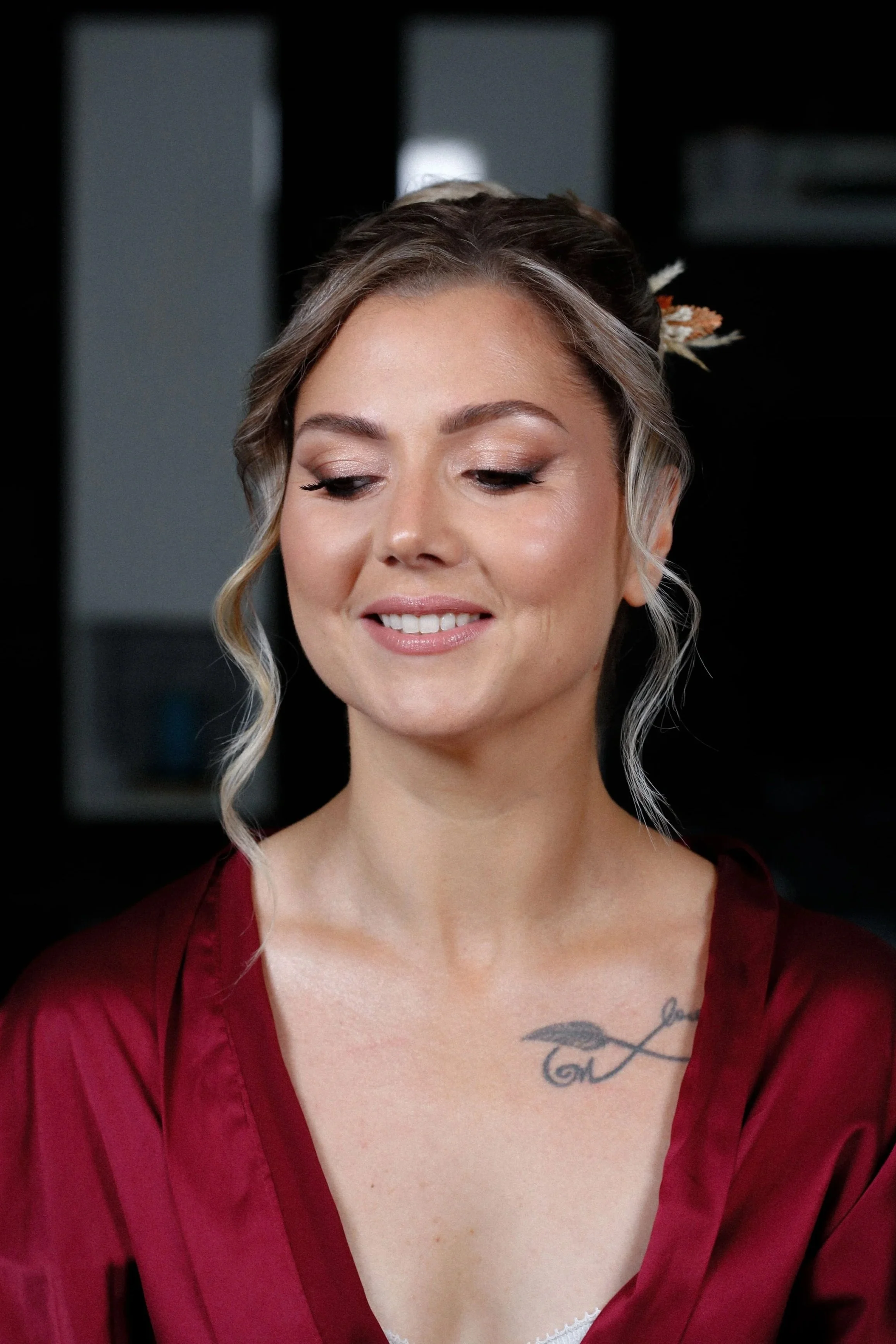 Une femme avec des cheveux coiffés, portant une robe rouge en satin, souriante, avec un tatouage d'une plume sur la poitrine, dans un intérieur sombre.