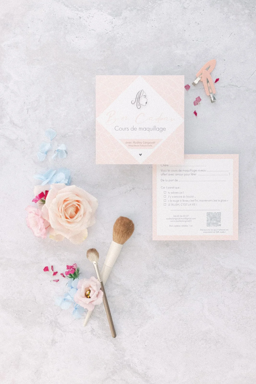 Cours de maquillage avec un pinceau, fleurs artisanales, cartes et petite figurine en bois, sur une surface claire.