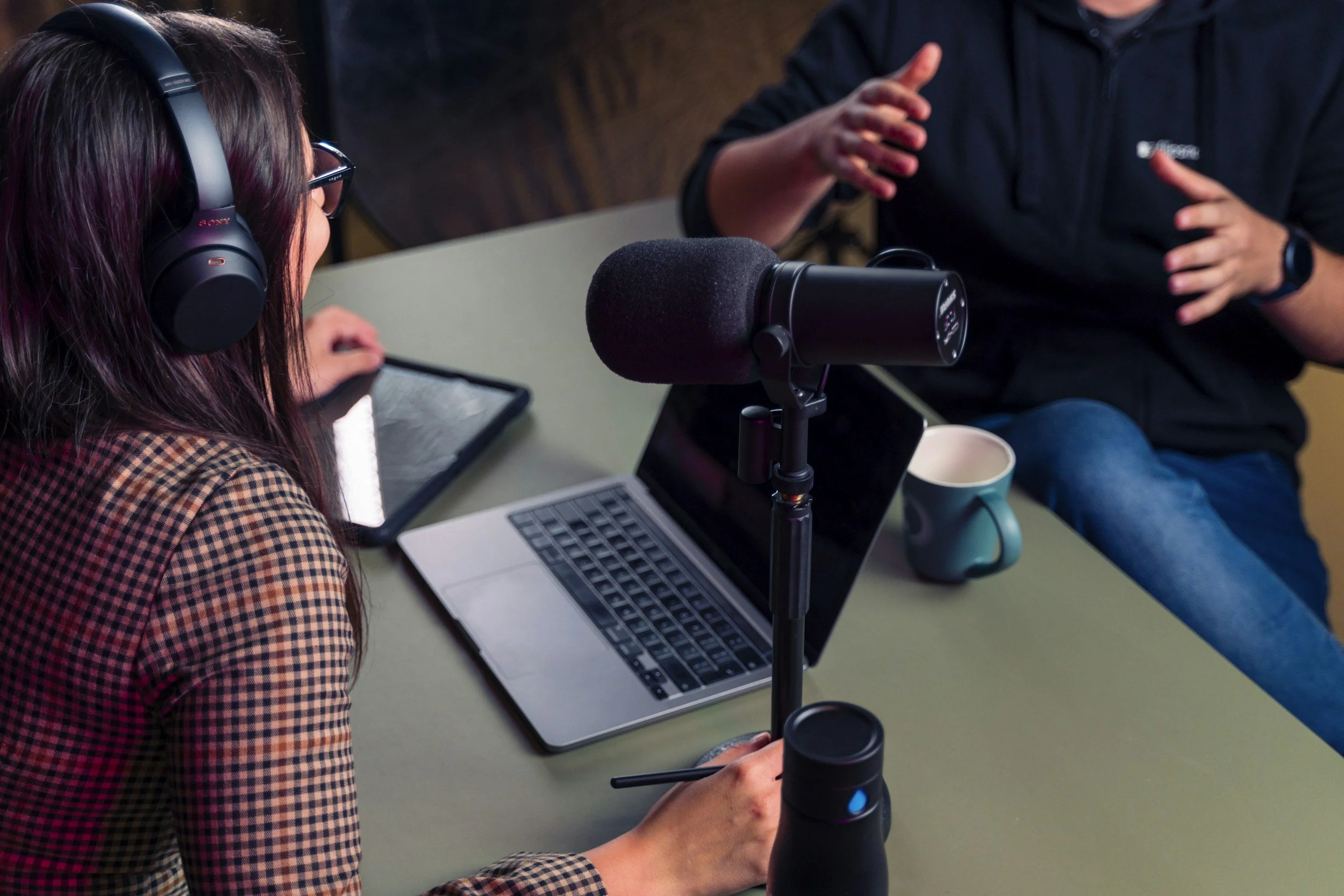 Two people recording a podcast at a table with a microphone, laptop, and tablet.